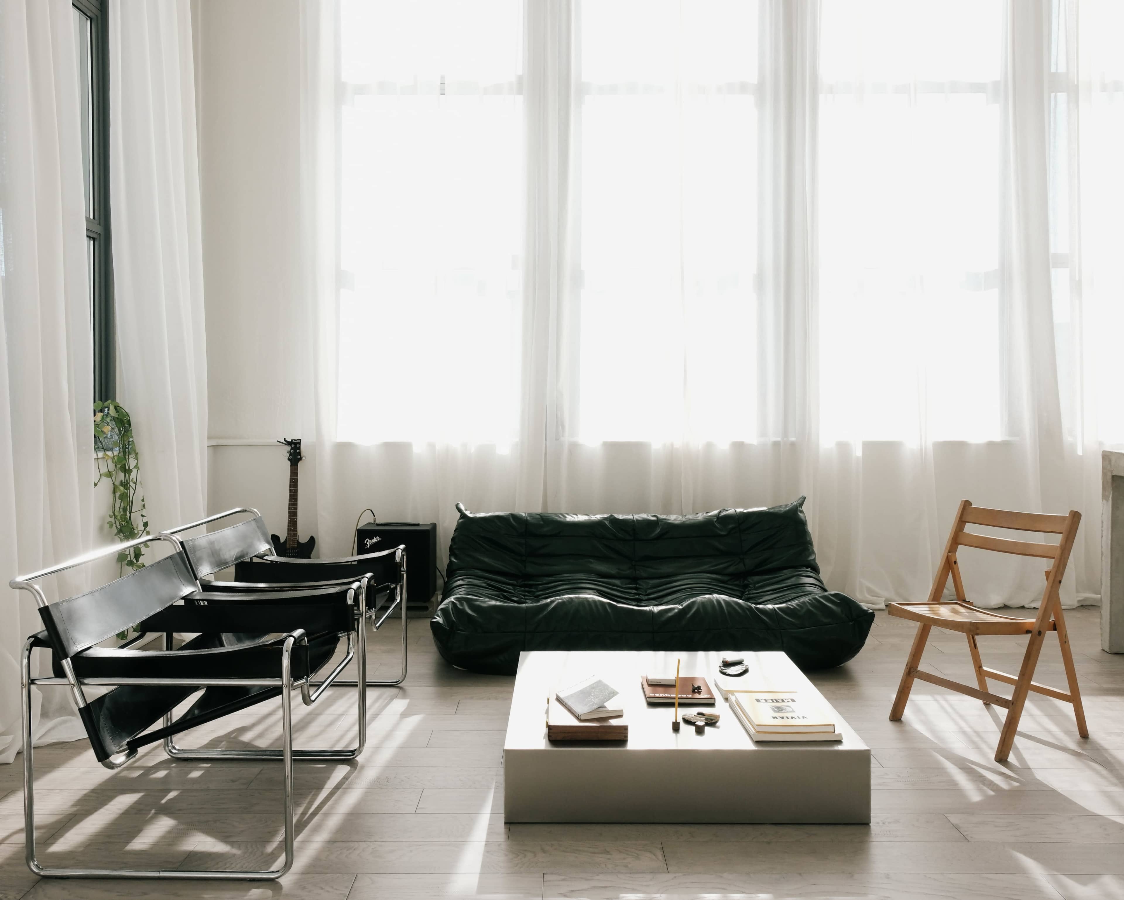 The image shows a modern living room with a black sofa, two black chairs, a wooden folding chair, and a coffee table holding books and small objects, all illuminated by natural light from large windows.
