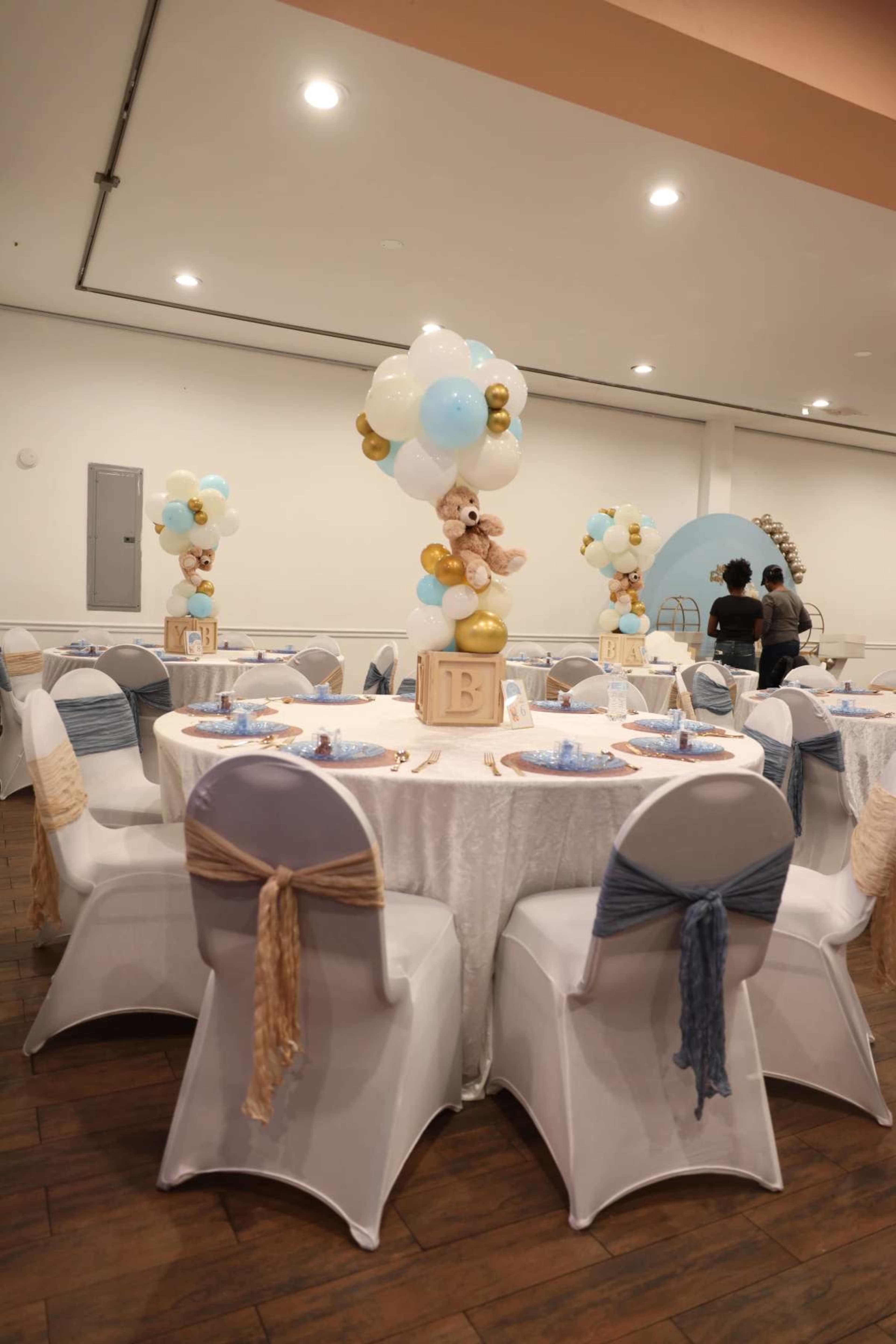 The image shows a decorated event space with round tables covered in white tablecloths, each featuring balloon centerpieces and place settings.