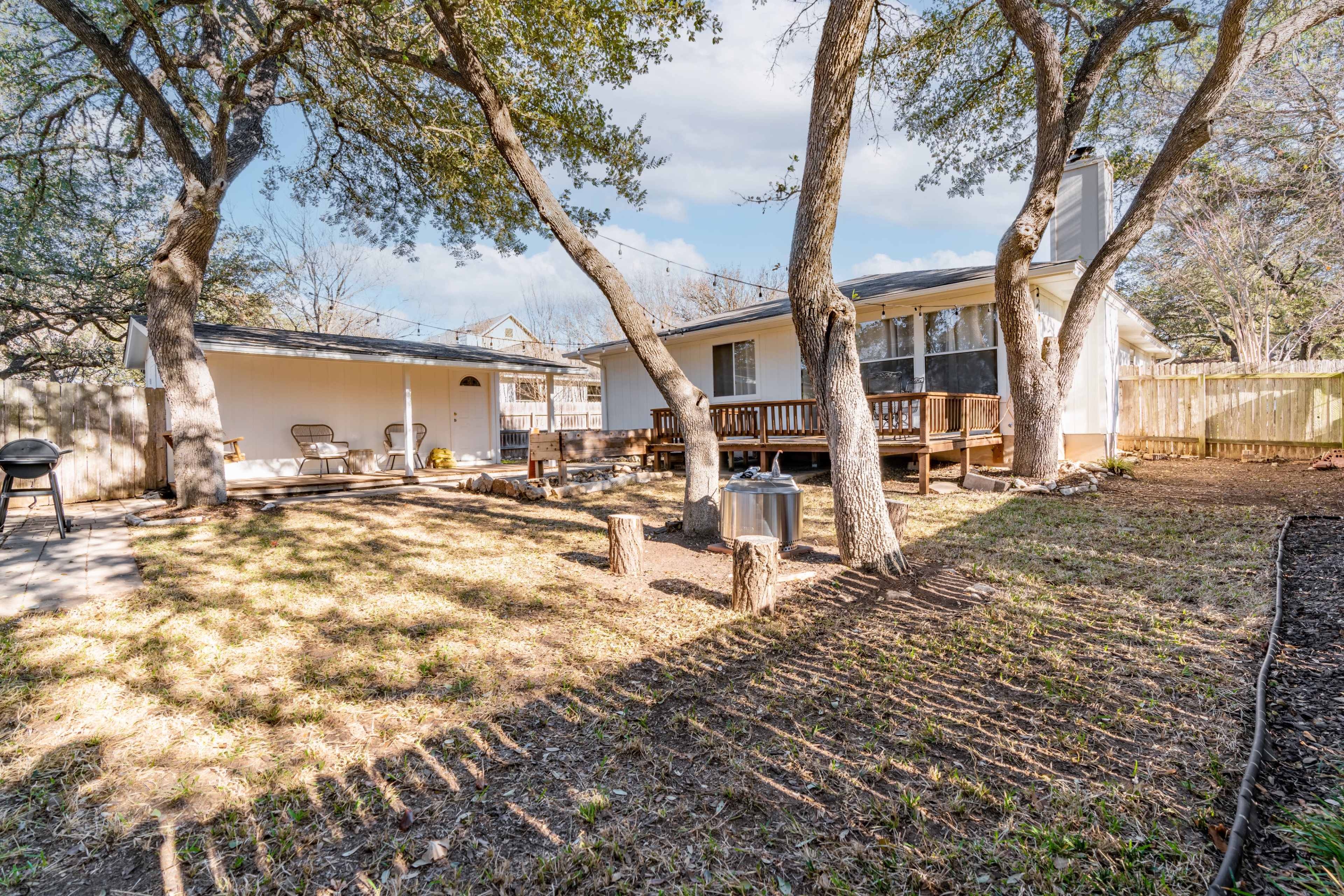 The image shows a serene backyard featuring two large trees, a wooden deck, and a patio area with outdoor furniture.