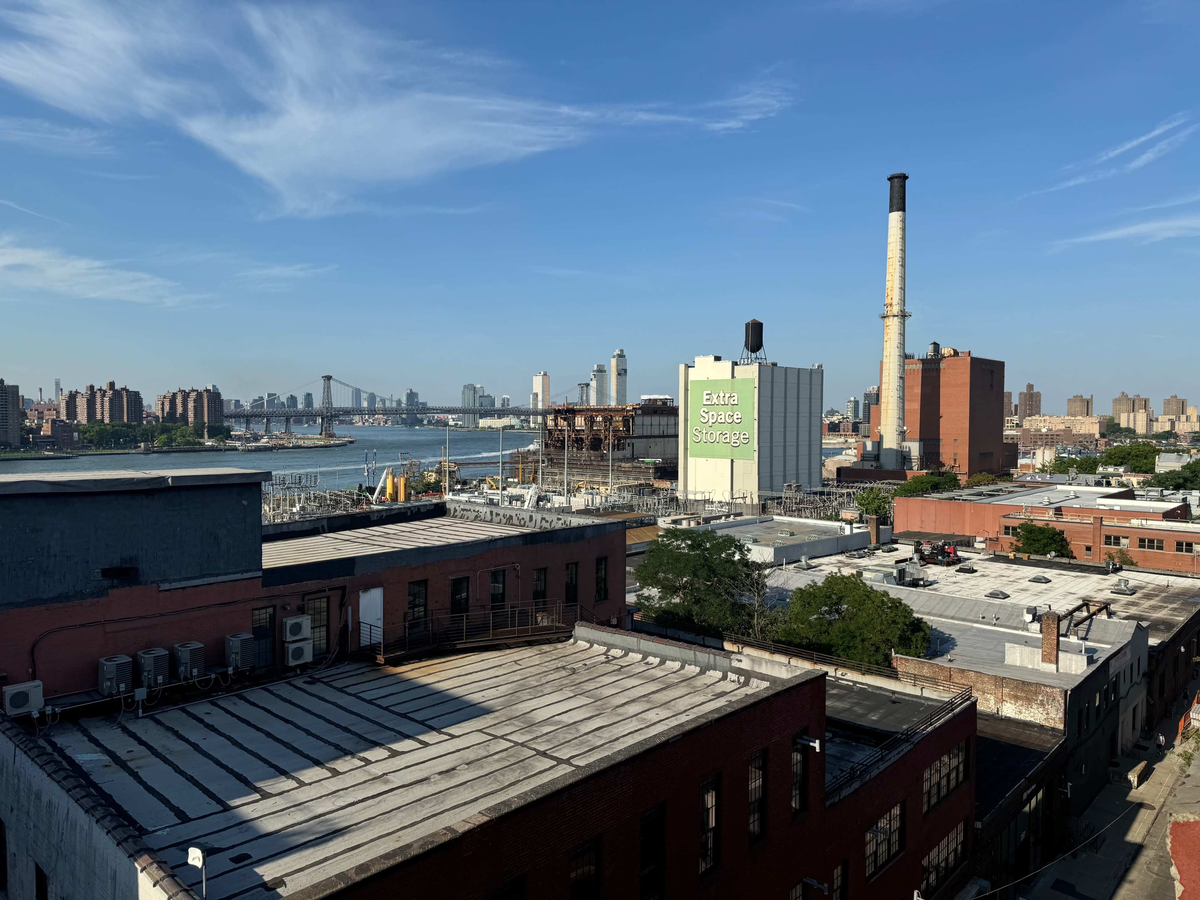 A city skyline with industrial buildings, a storage facility, and a bridge in the background, under a clear blue sky.