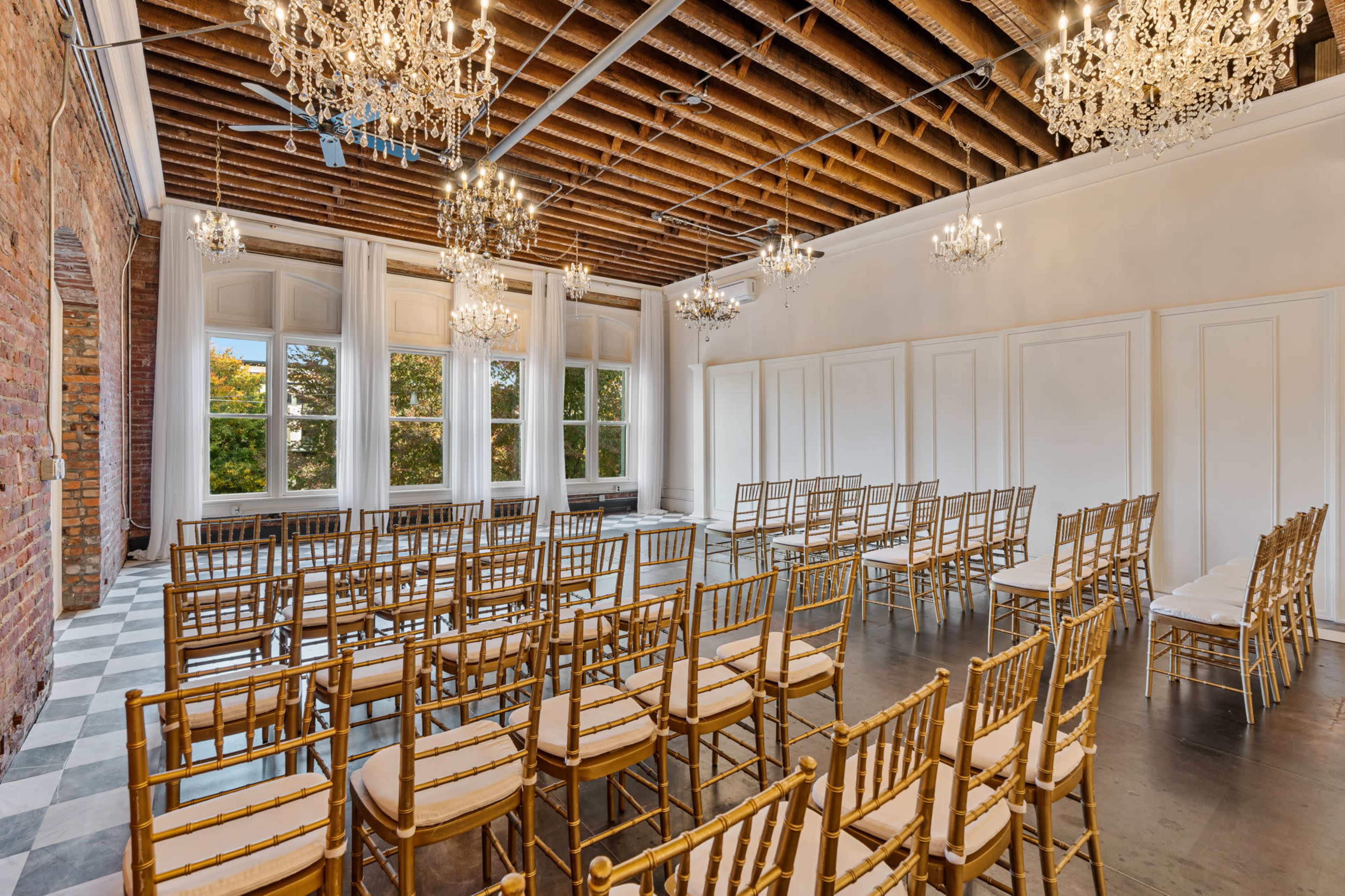 The image shows an elegant room set up for a formal event, featuring rows of gold chiavari chairs, chandeliers, and large windows with natural light.