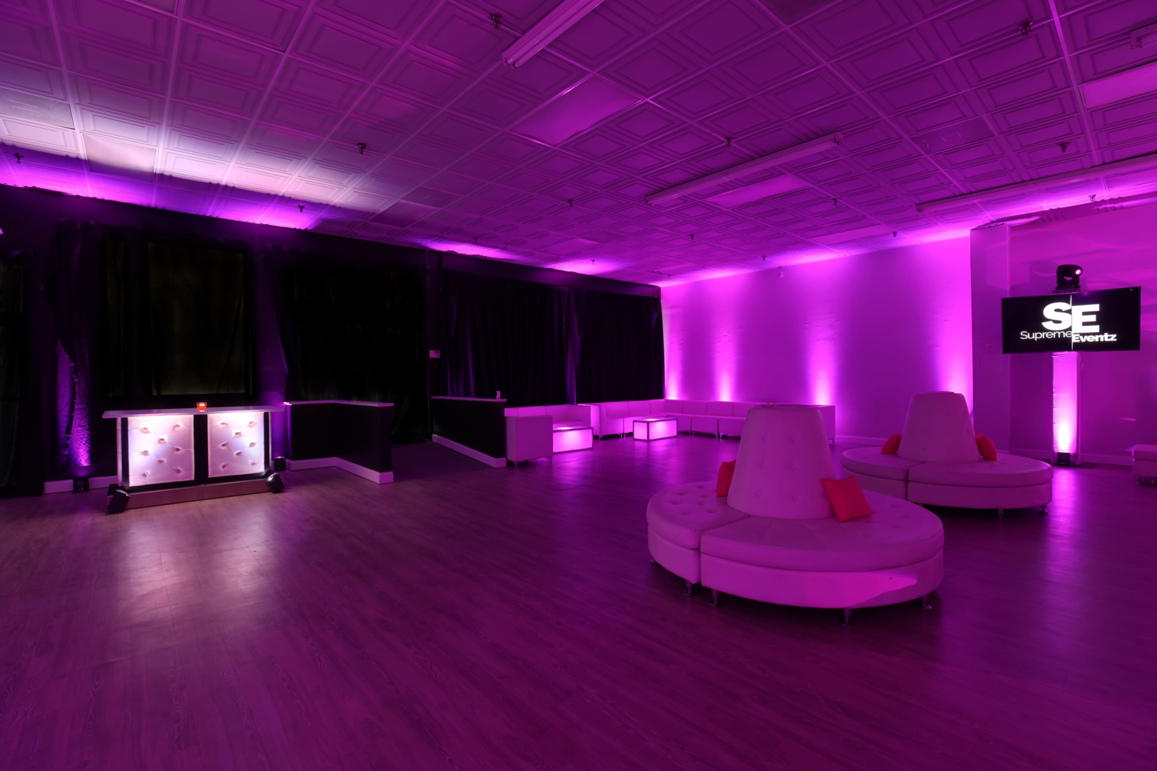 The room features a spacious layout with purple lighting, circular white seating, and a simple bar setup against a black backdrop.