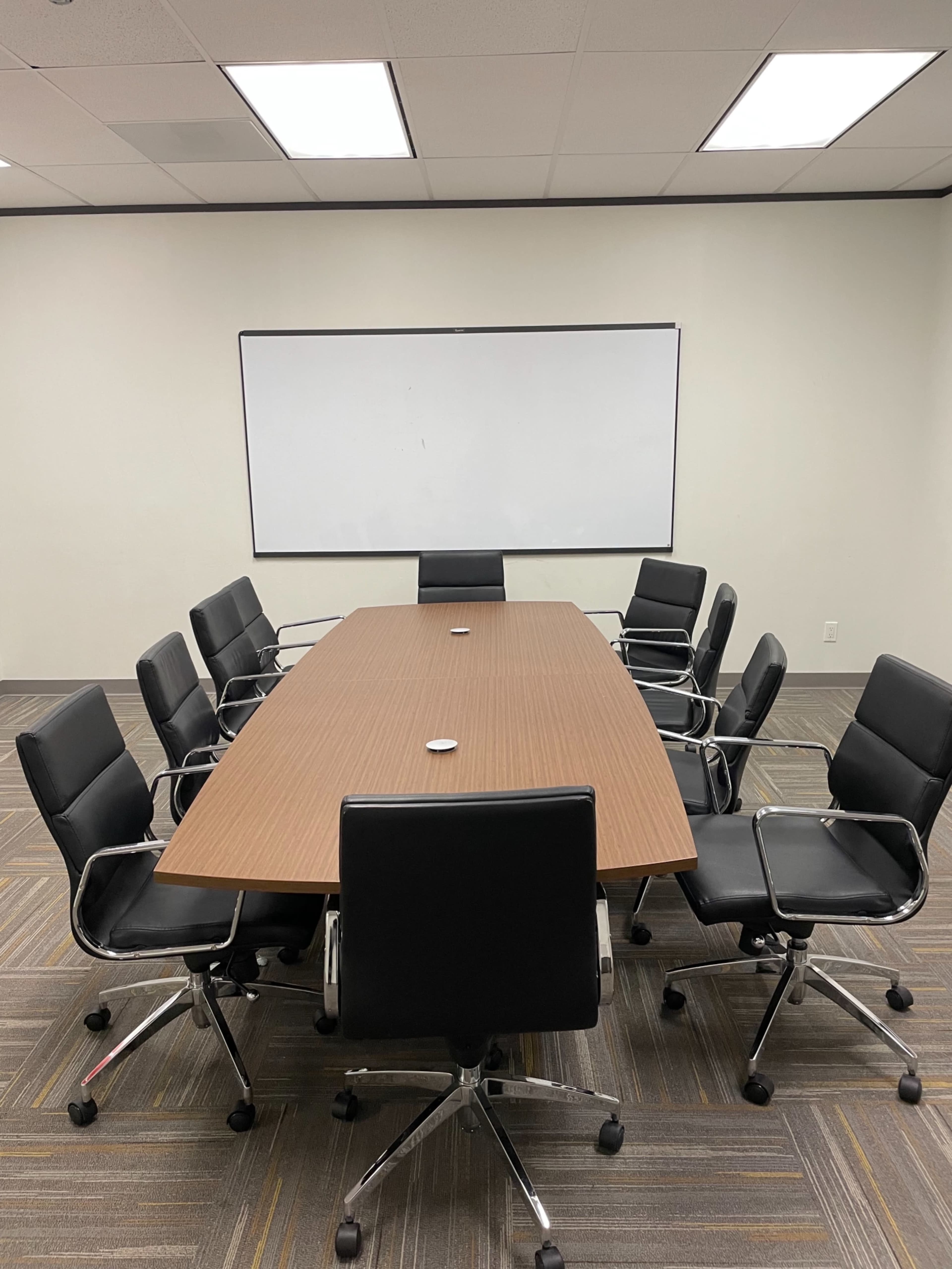 A conference room features a long wooden table surrounded by eight black rolling chairs and a whiteboard on the wall.
