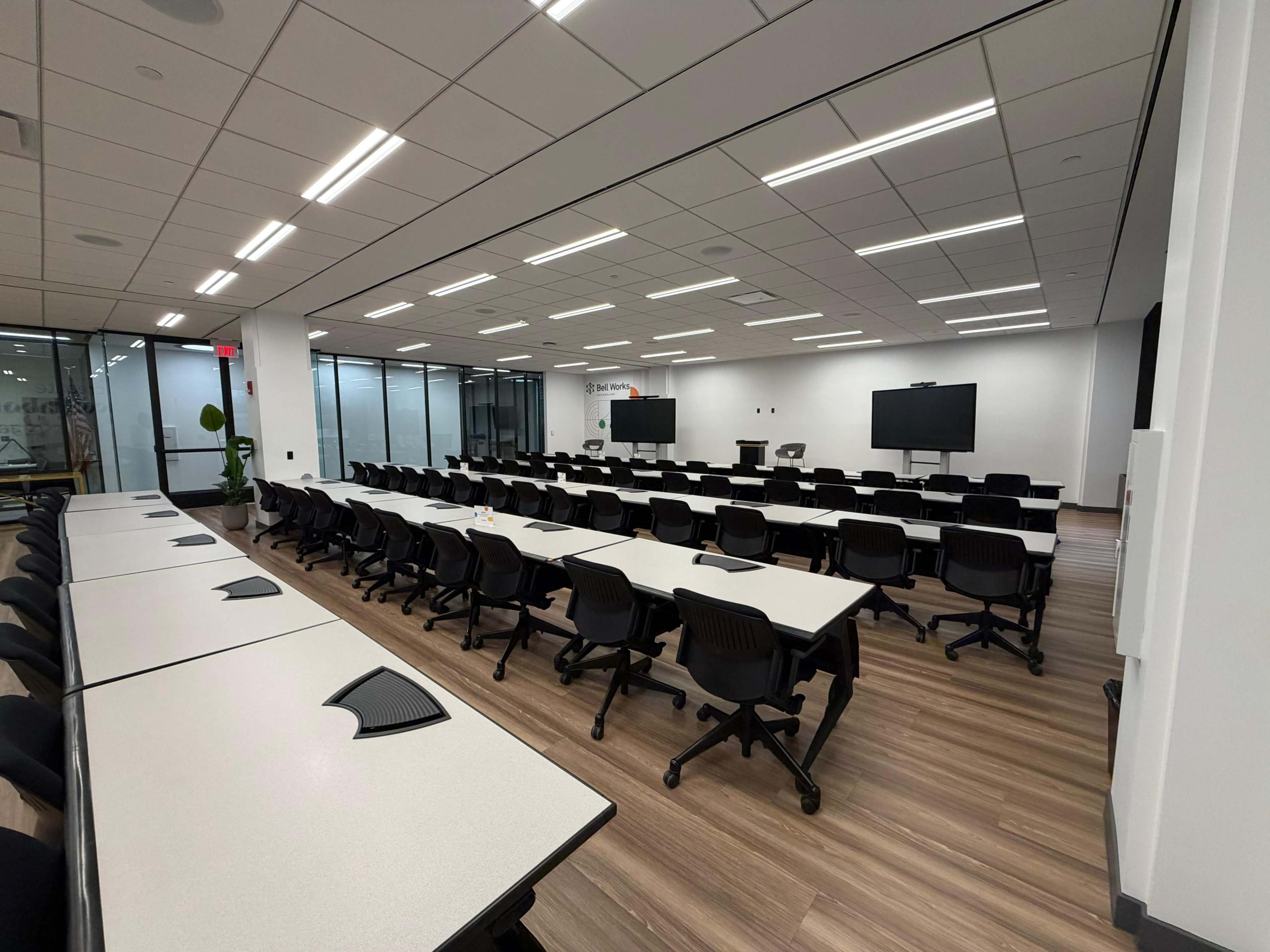 The image shows a spacious conference room arranged with rows of tables and chairs, featuring large screens and glass walls.