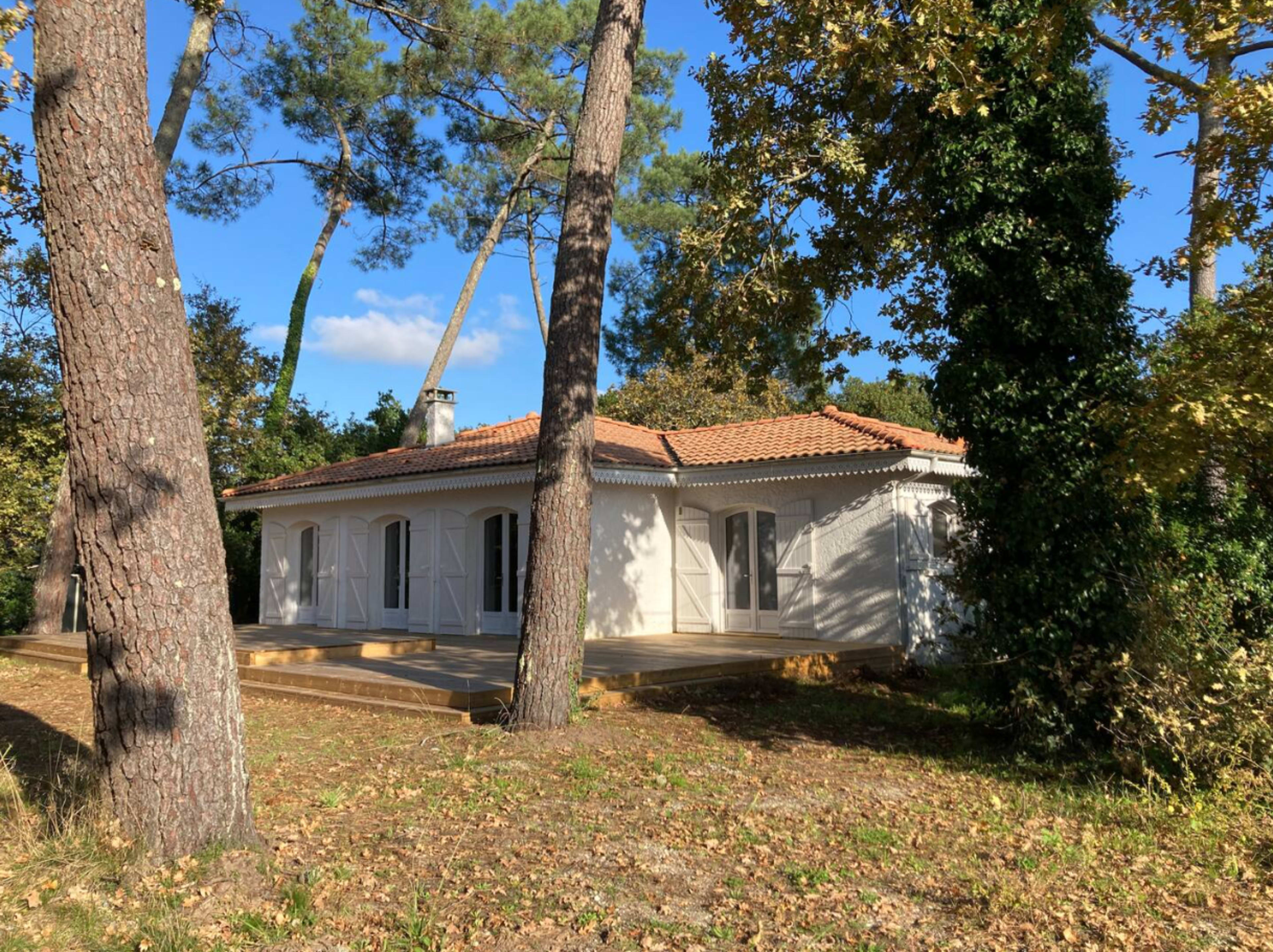 A single-story house with white exterior walls and a red-tiled roof is set among tall pine trees and surrounded by greenery.
