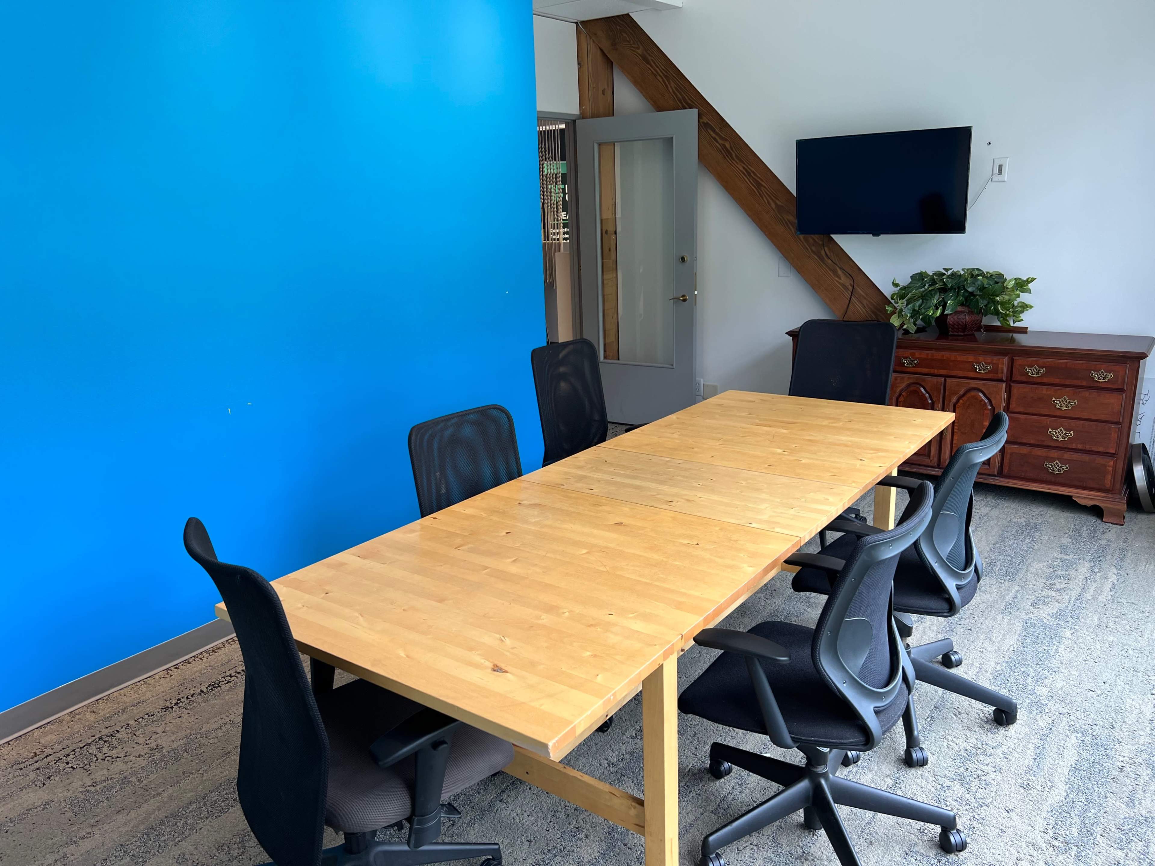 A large wooden table surrounded by black office chairs is positioned in a brightly lit meeting room with a blue wall.