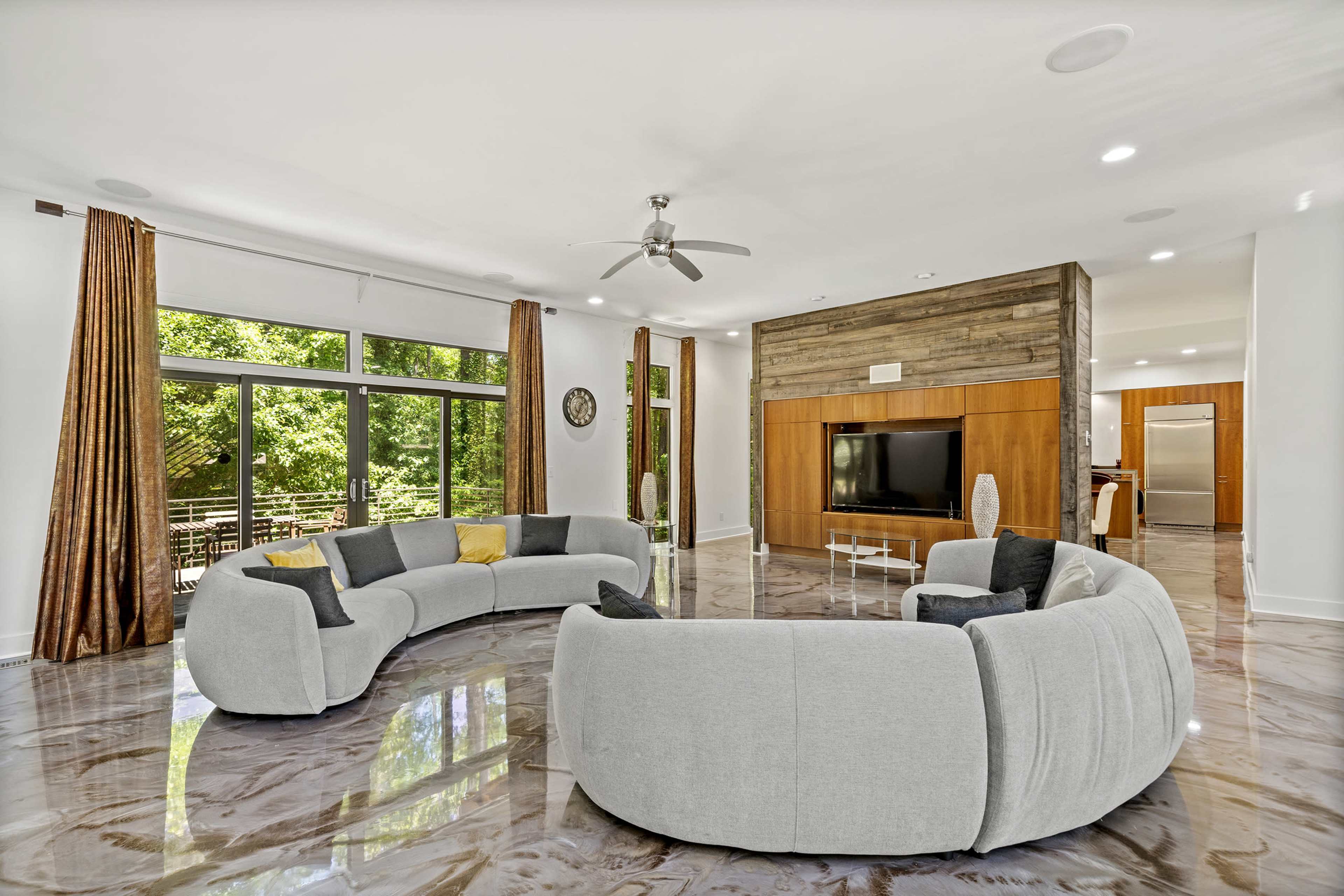 The image shows a modern living room featuring a curved gray sofa, large windows with natural light, and a wooden accent wall housing a television.