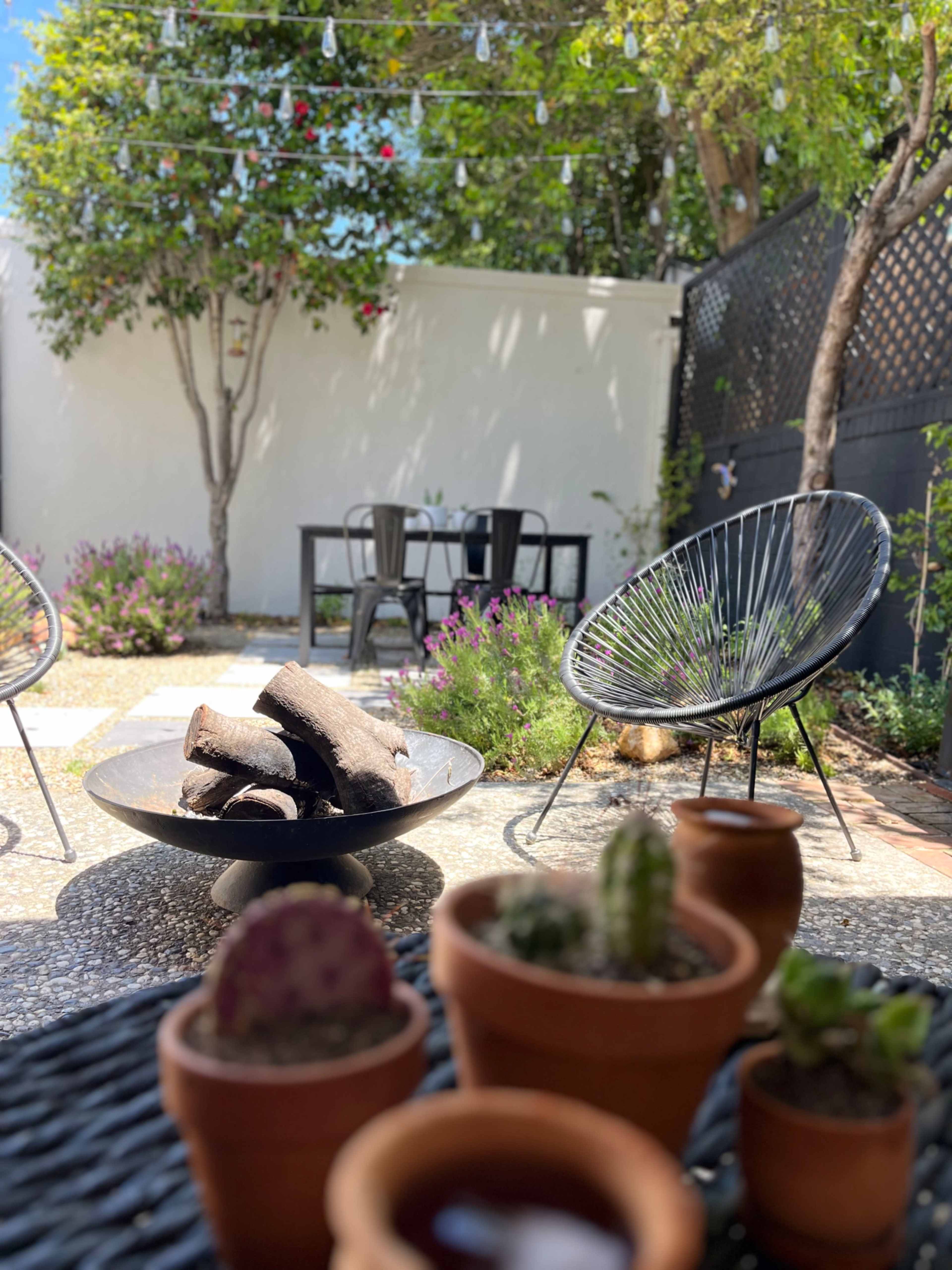 A patio scene features a fire pit surrounded by modern chairs, with a table in the background and potted plants in the foreground.