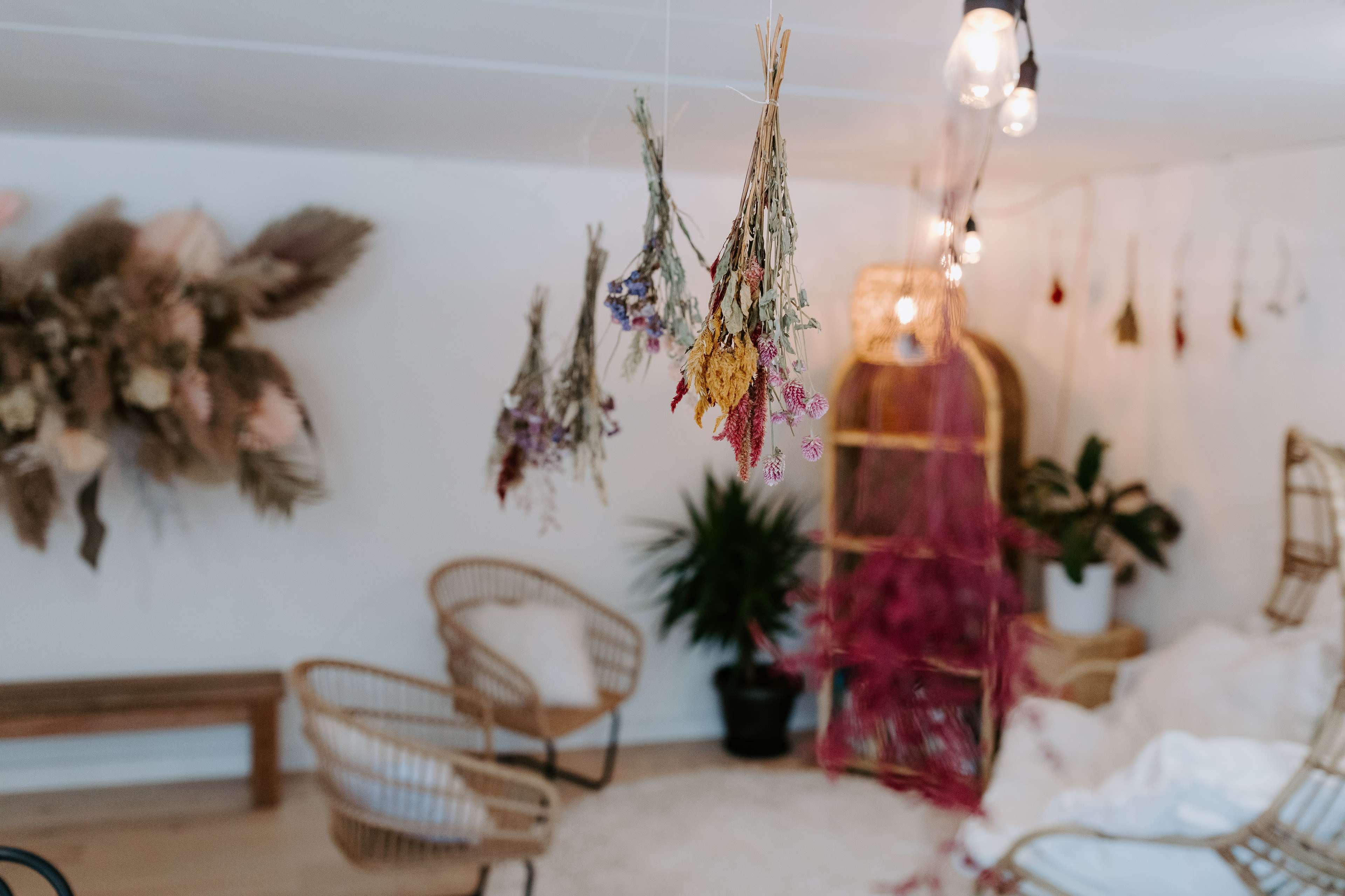 The image shows a cozy room decorated with hanging dried flowers, bamboo chairs, and soft textiles, featuring a plant and warm lighting.