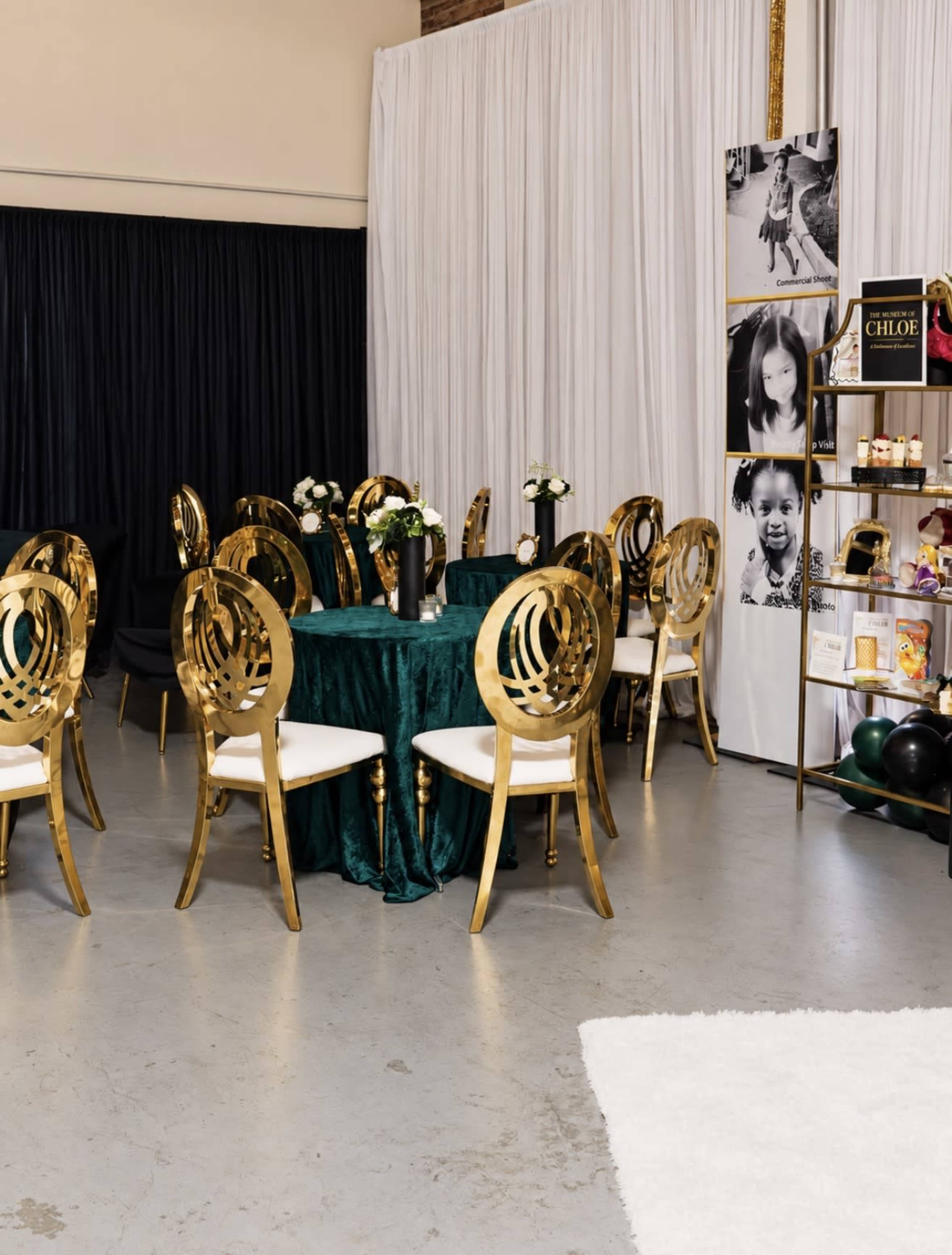 The scene features a decorated event space with gold chairs around a green table, white drapes in the background, and a display wall with photographs and decorative items.