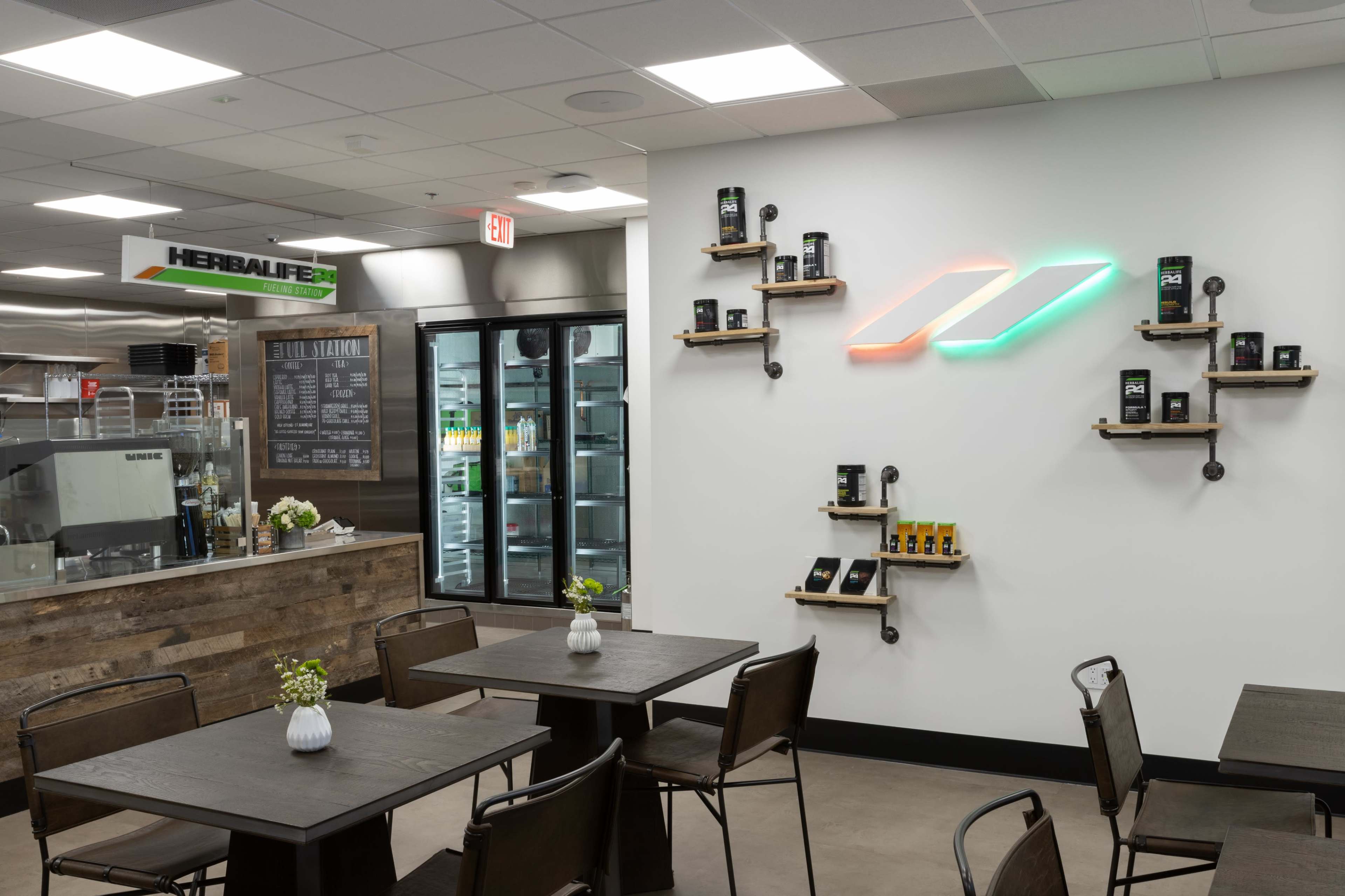 The image shows a modern café interior with tables and a wall displaying various Herbalife products on shelves.