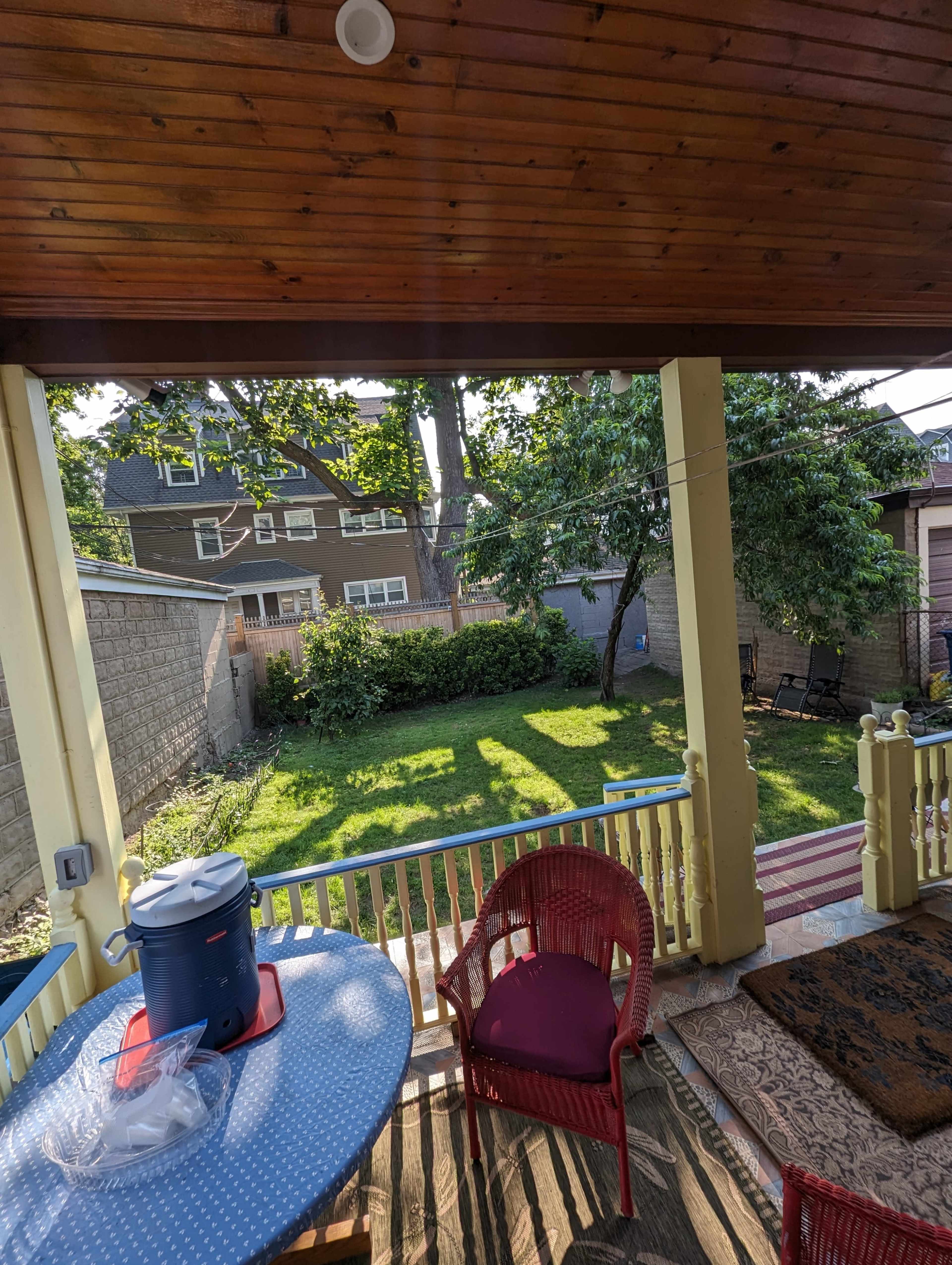 The image shows a back porch with a red chair and a table overlooking a green backyard featuring trees and a modest garden space.