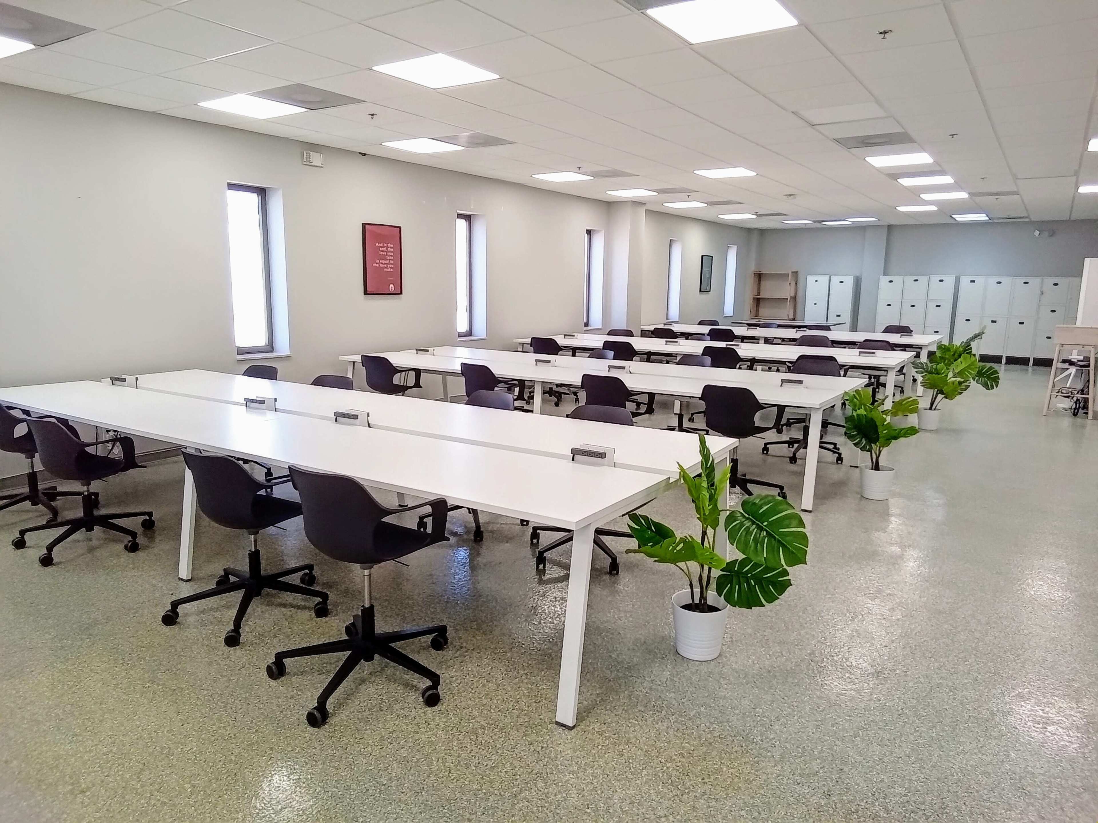 A spacious, modern office area with several long tables and rolling chairs, featuring potted plants and storage lockers along the wall.