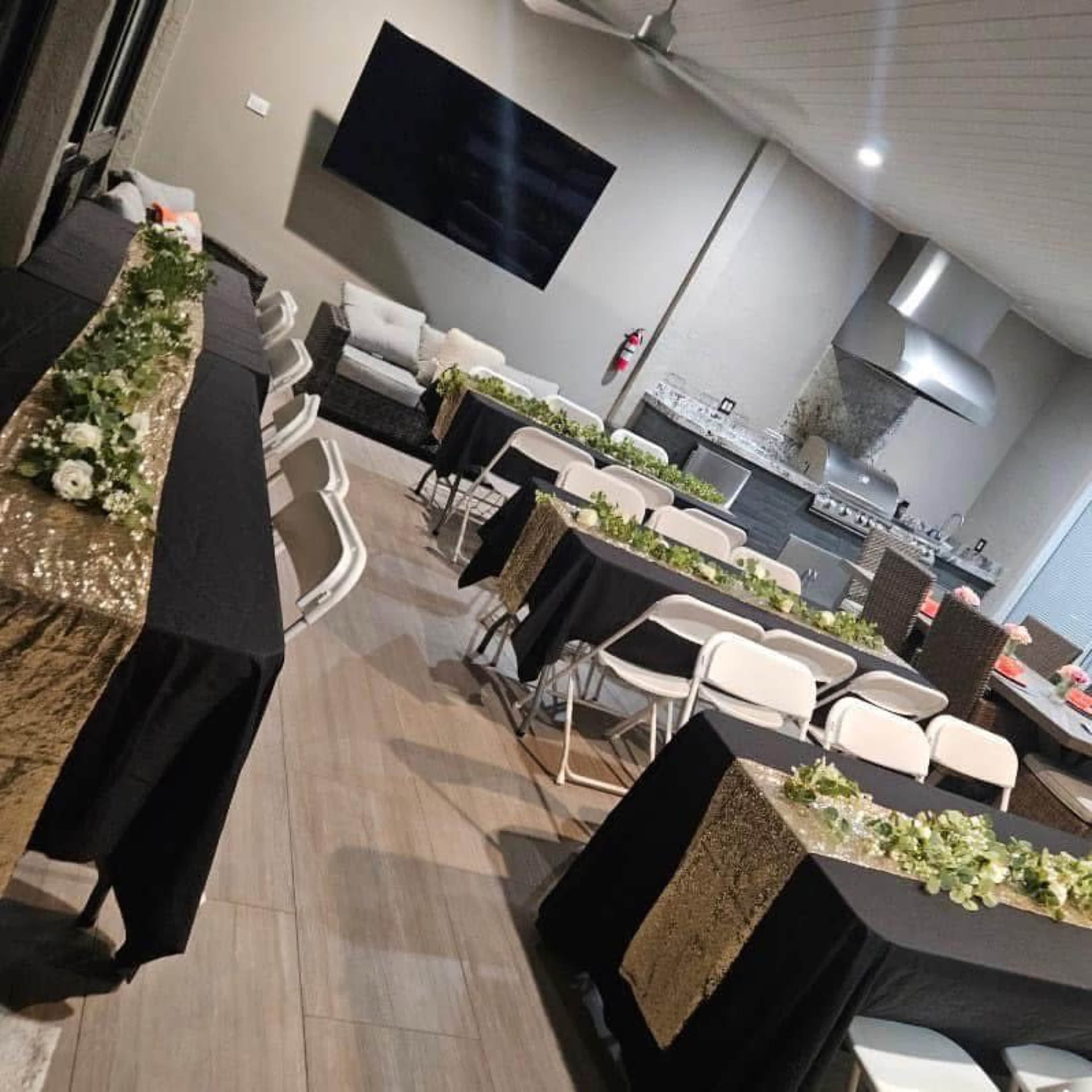 A spacious indoor event area features multiple tables covered with black tablecloths and adorned with gold sequin runners, surrounded by white chairs, and a large television mounted on the wall.