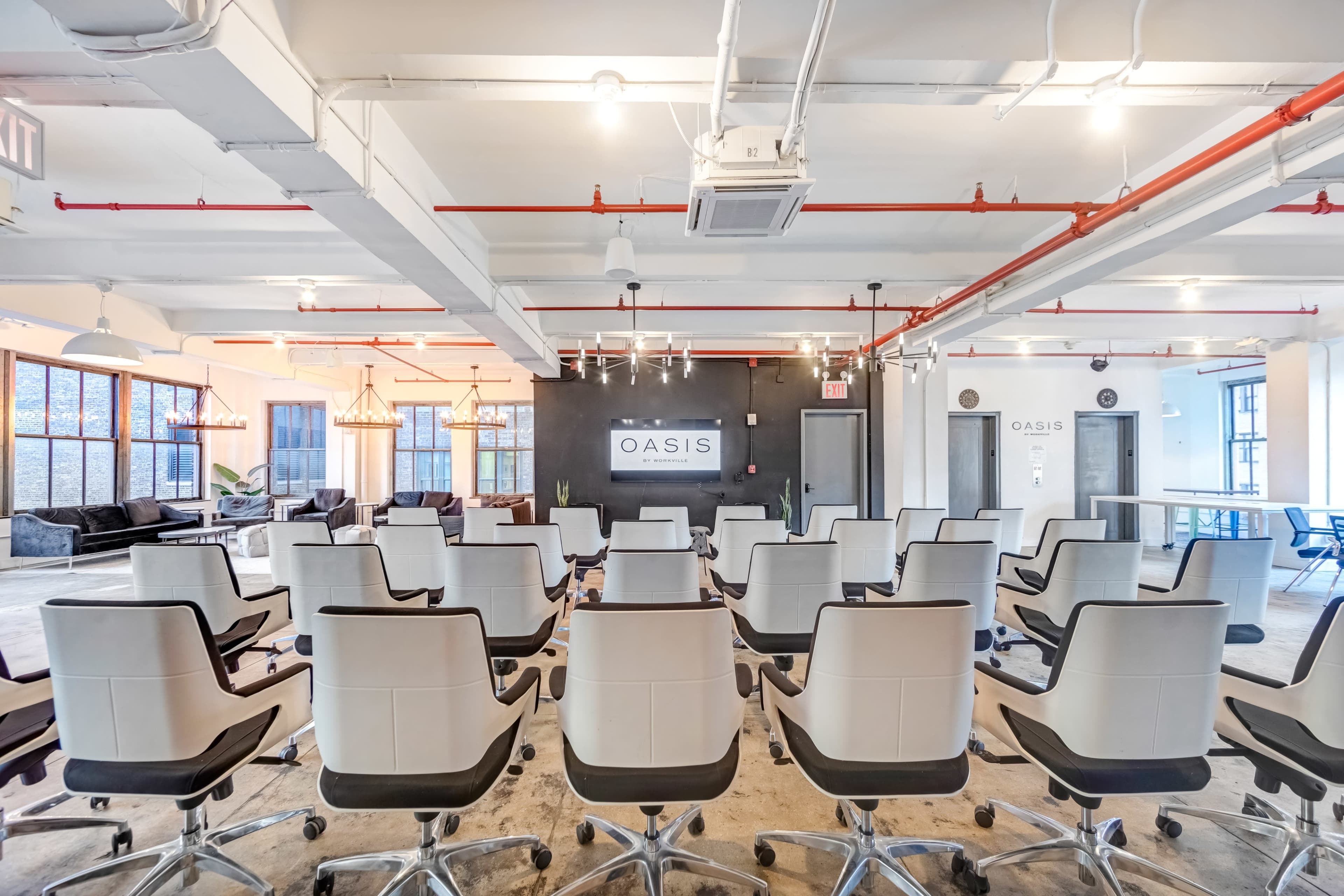A spacious meeting room with rows of white office chairs facing a presentation area, featuring large windows and modern lighting fixtures.