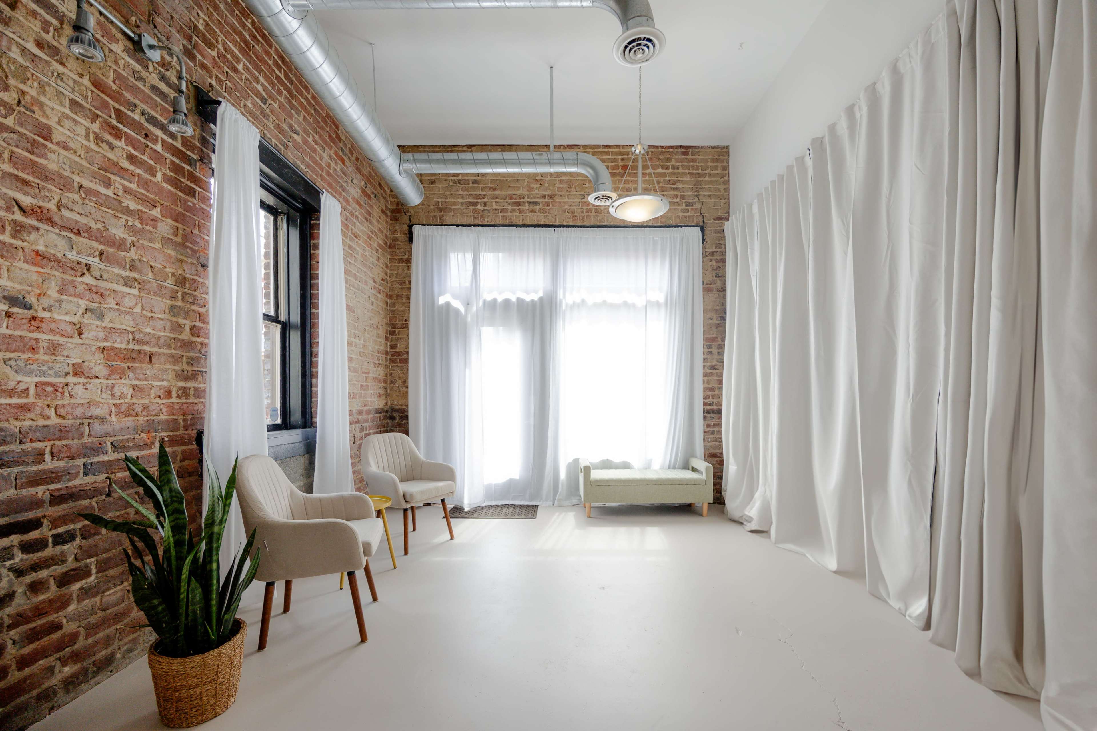 A spacious room with exposed brick walls, large windows covered by white curtains, two upholstered chairs, and a potted plant in the corner.