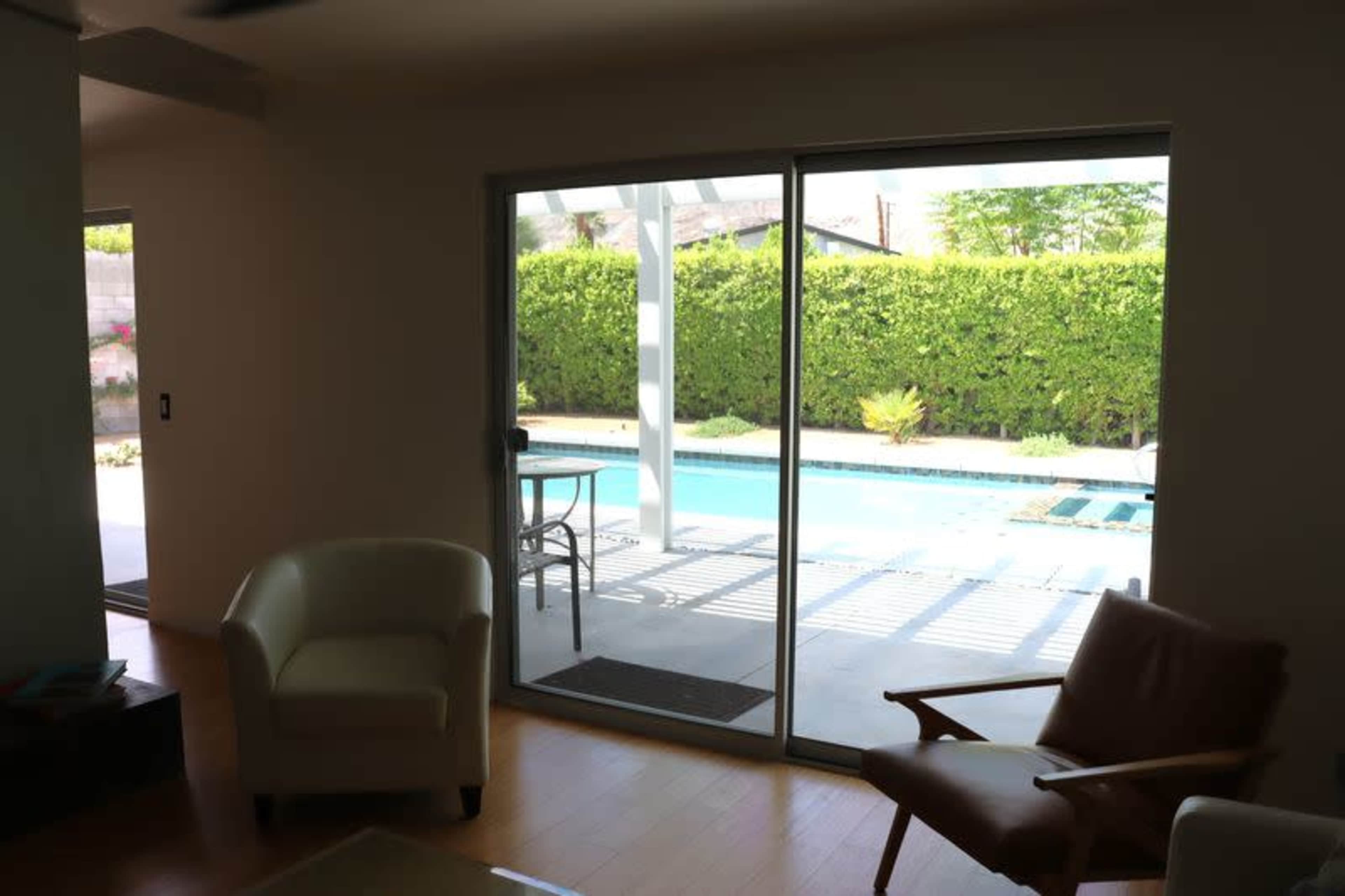 A living room features two chairs and a glass sliding door leading to a pool area surrounded by hedges.