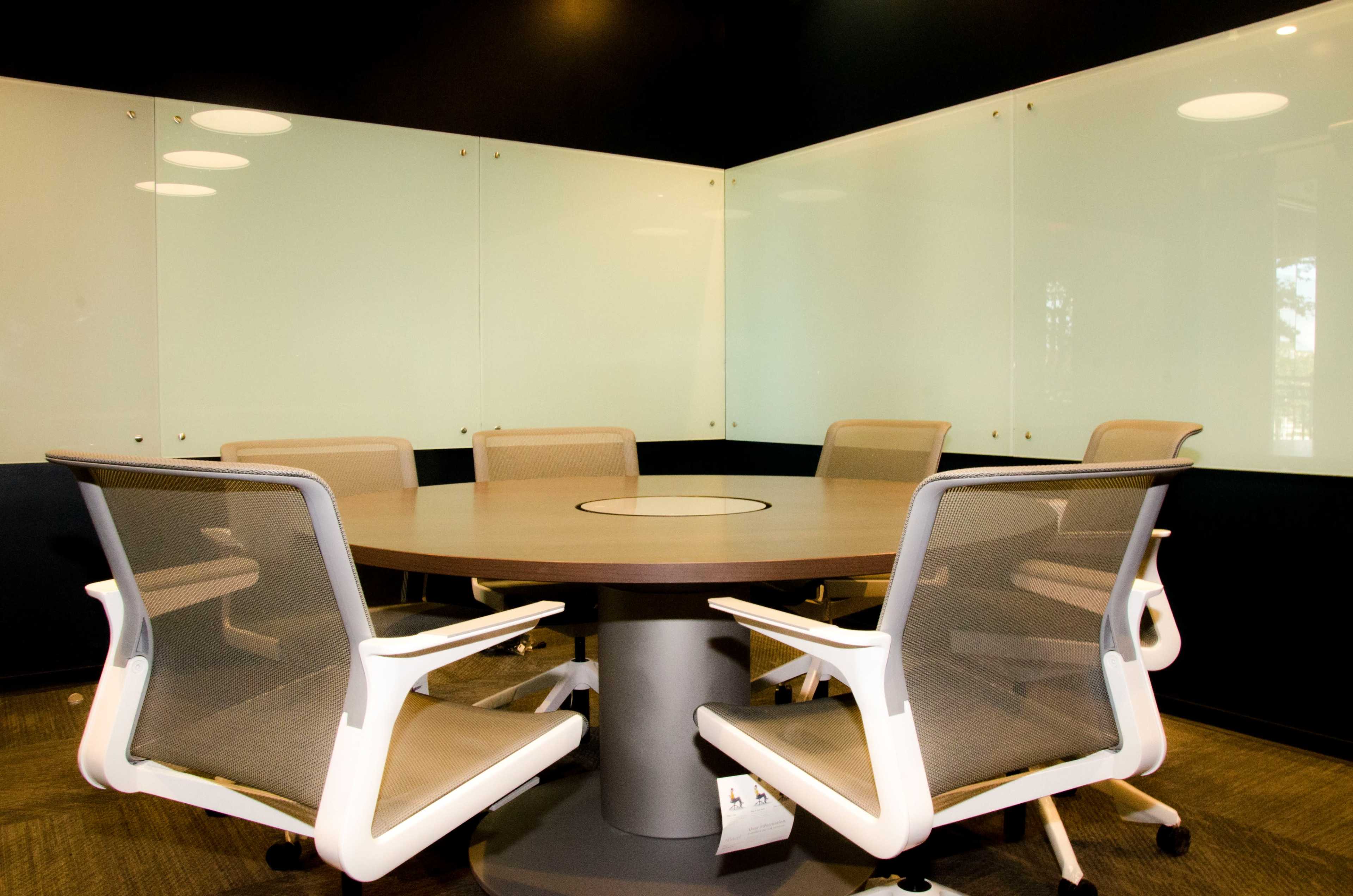A conference room features a round table surrounded by six ergonomic chairs and glass walls.