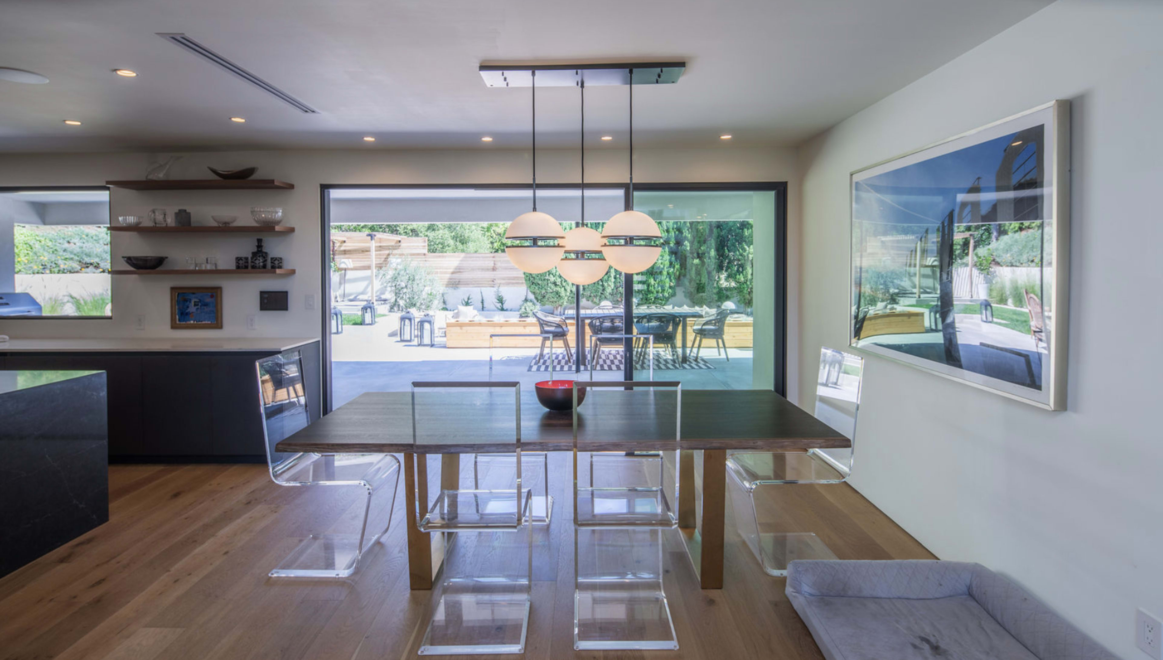 A modern dining area features a wooden table surrounded by transparent chairs, with a view of an outdoor patio through large glass doors.