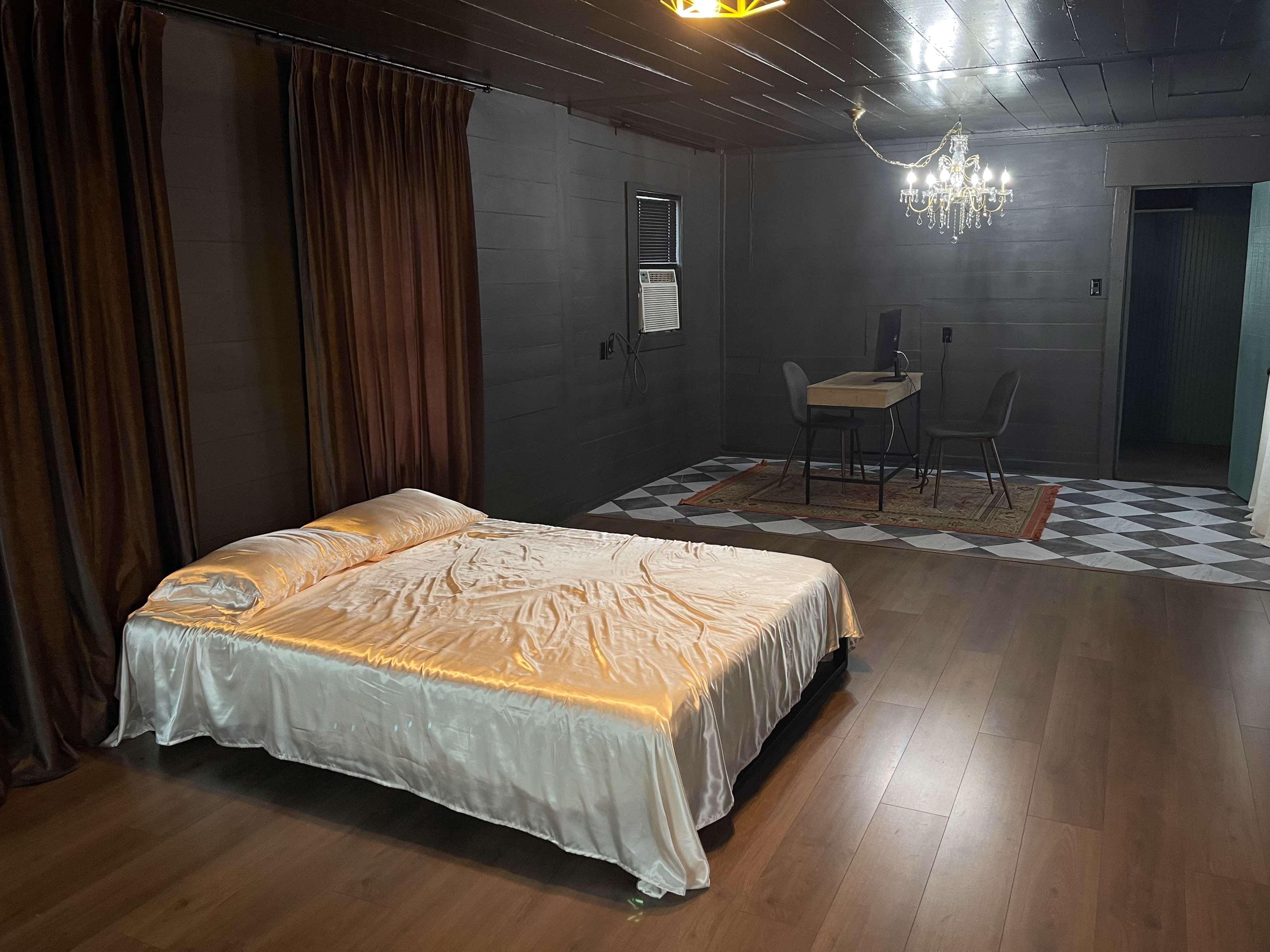 The image shows a sparsely furnished room with a bed covered in a shiny white bedspread, a small dining table with chairs, and a chandelier overhead.