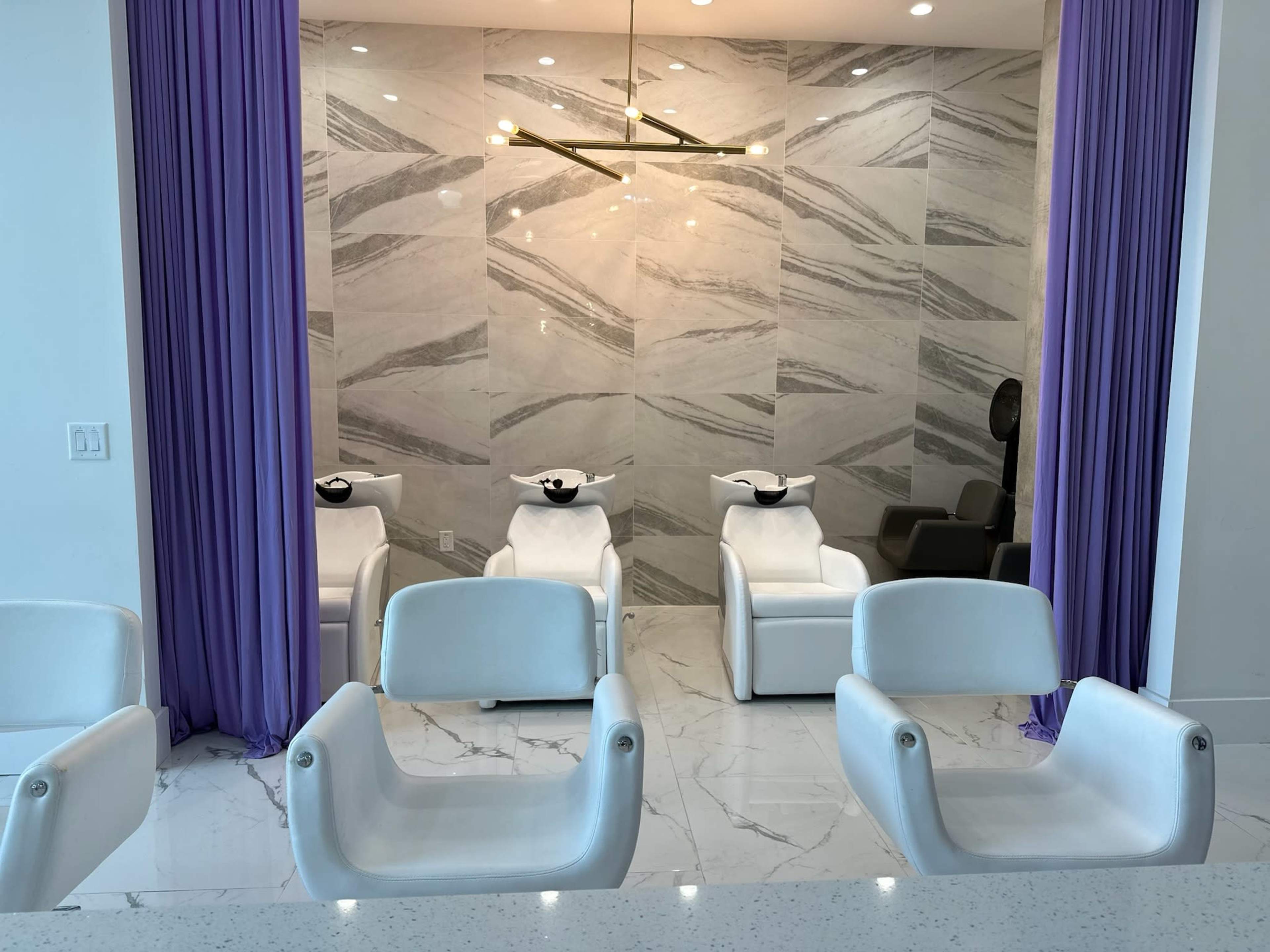 The image shows a modern hair salon with three reclining shampoo chairs positioned against a marble wall, separated by purple curtains.