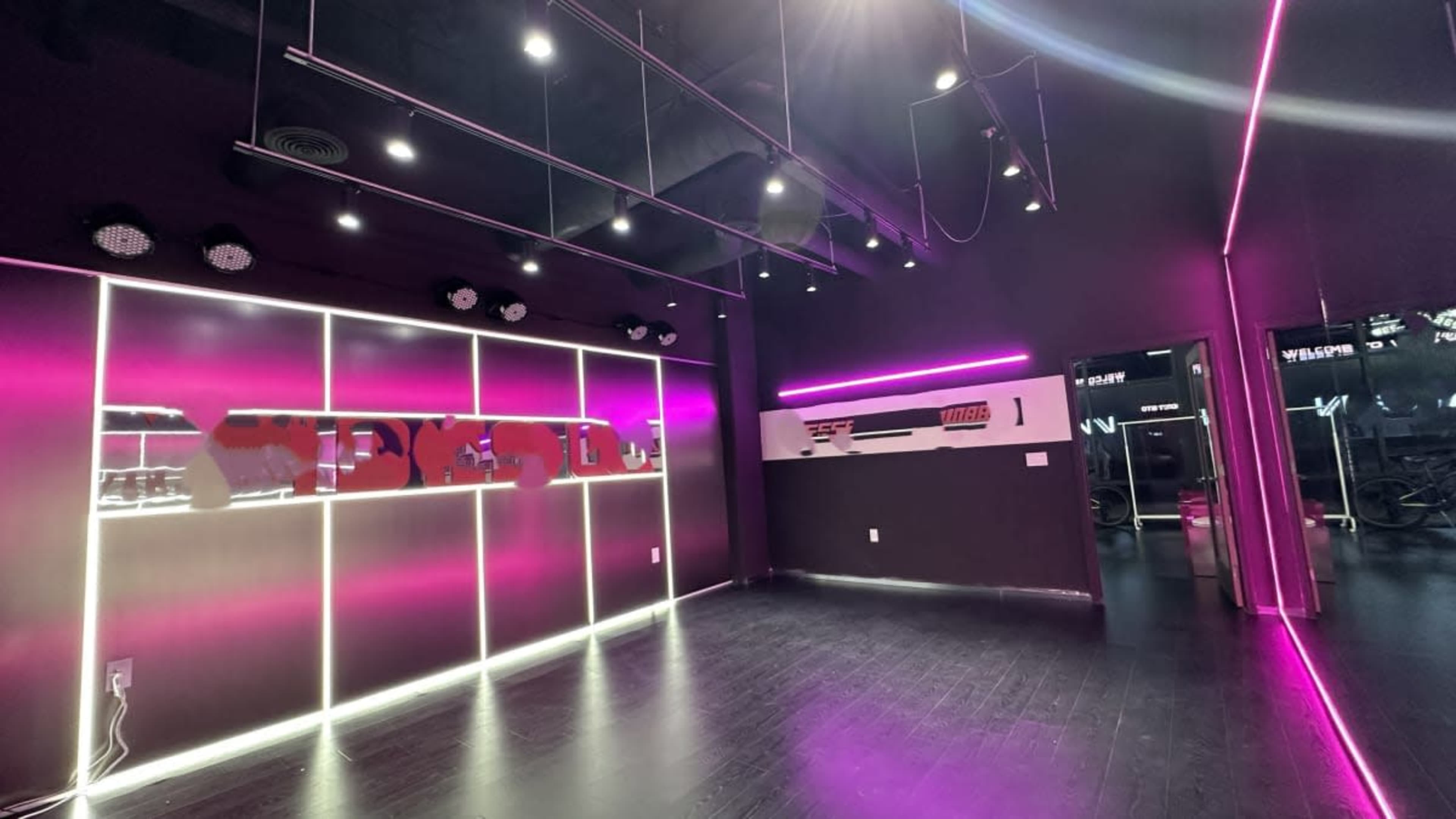 The image shows a modern fitness studio with black walls, pink neon lighting, and a mirrored wall.