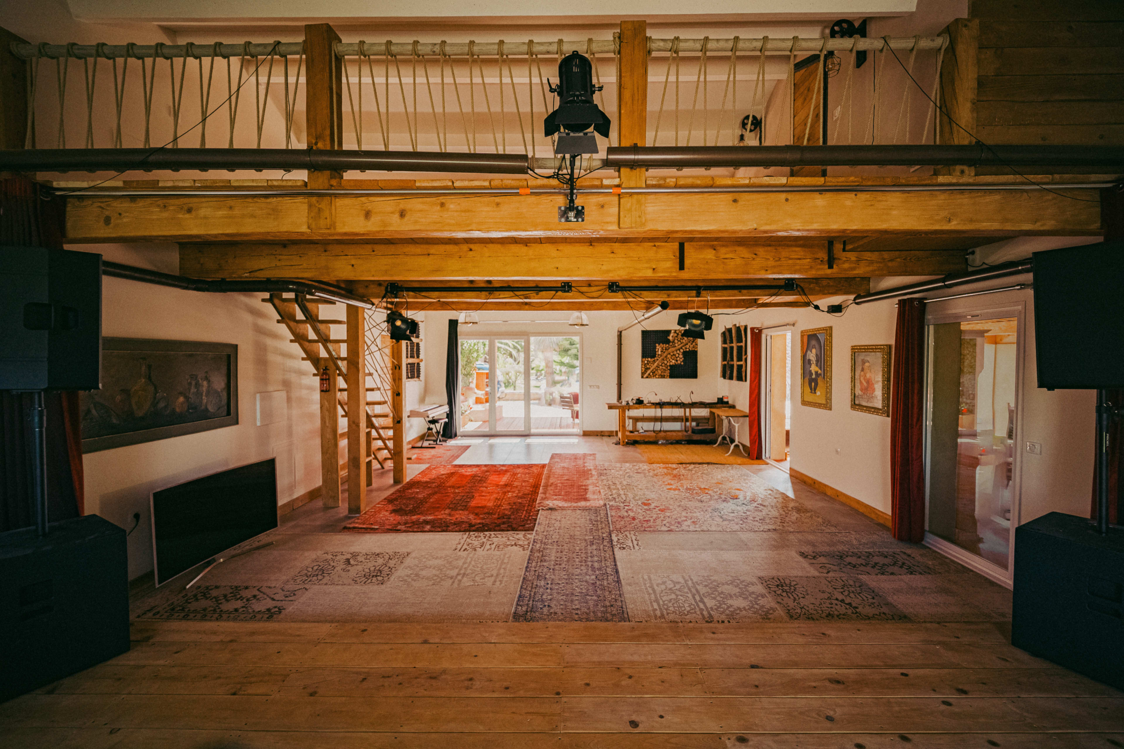 The image shows a spacious interior with wooden beams, a staircase, and area rugs on the floor, leading to large glass doors that open to an outdoor area.