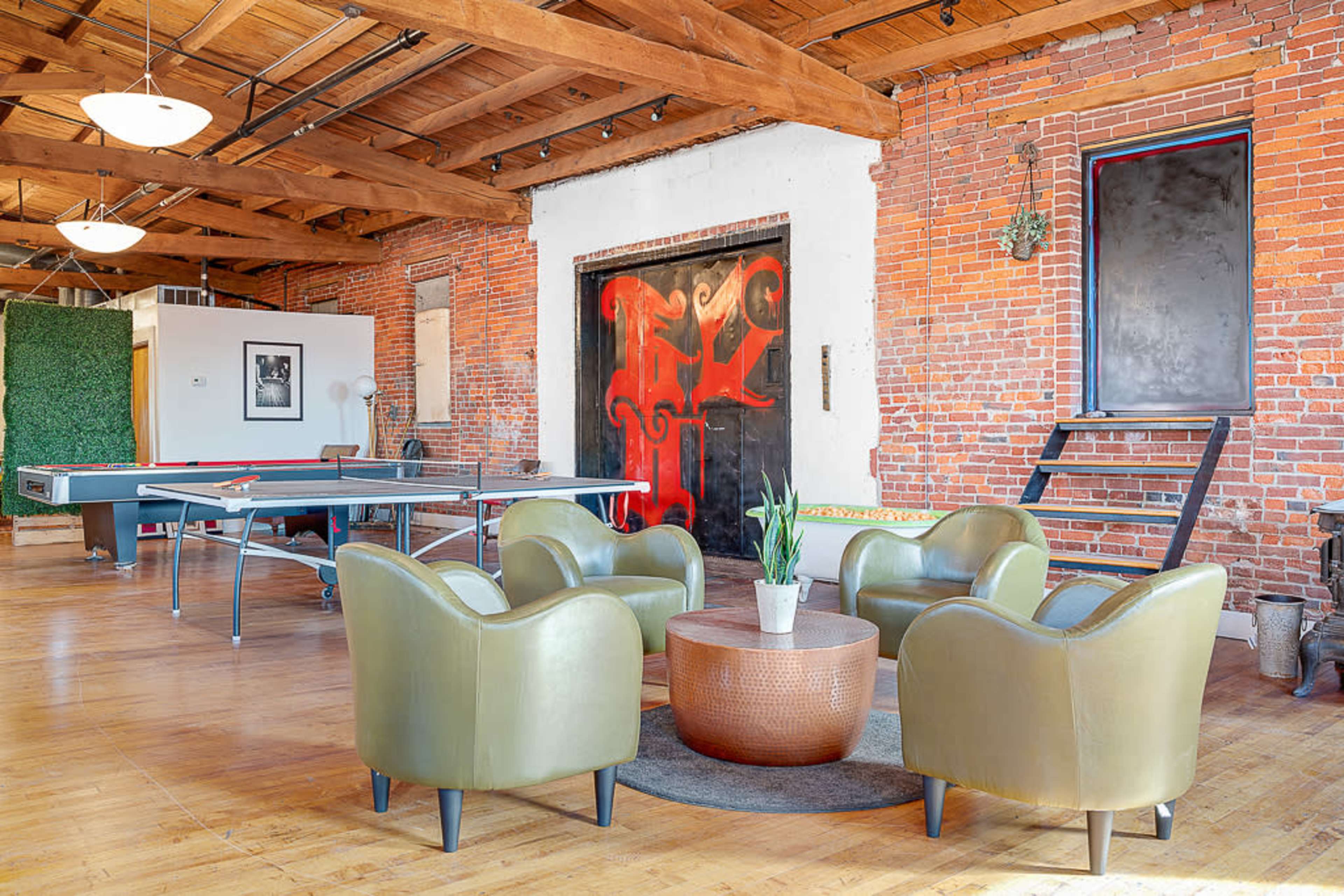 A spacious, industrial-style lounge area featuring a table tennis setup and four green chairs arranged around a round coffee table on a wooden floor.