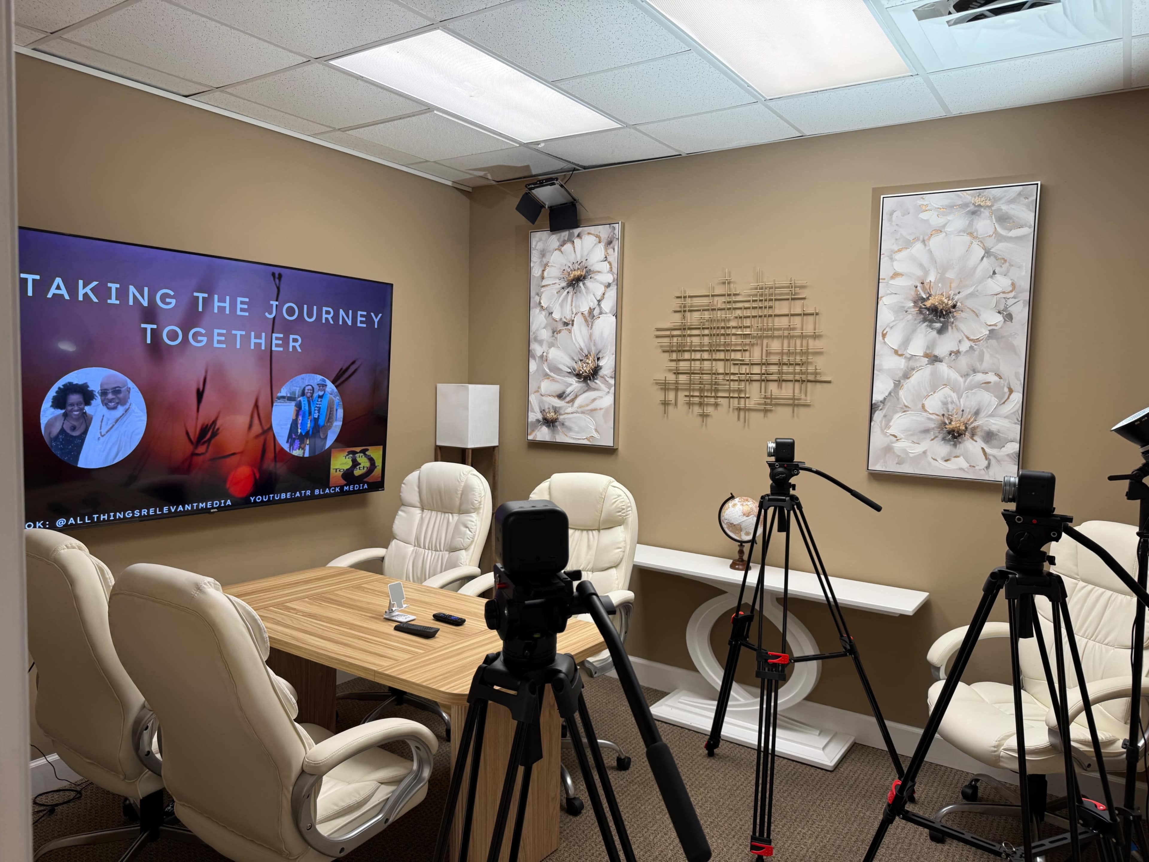 The image shows a modern conference room equipped with a large screen for presentations, a wooden table surrounded by white chairs, and several cameras positioned for filming.