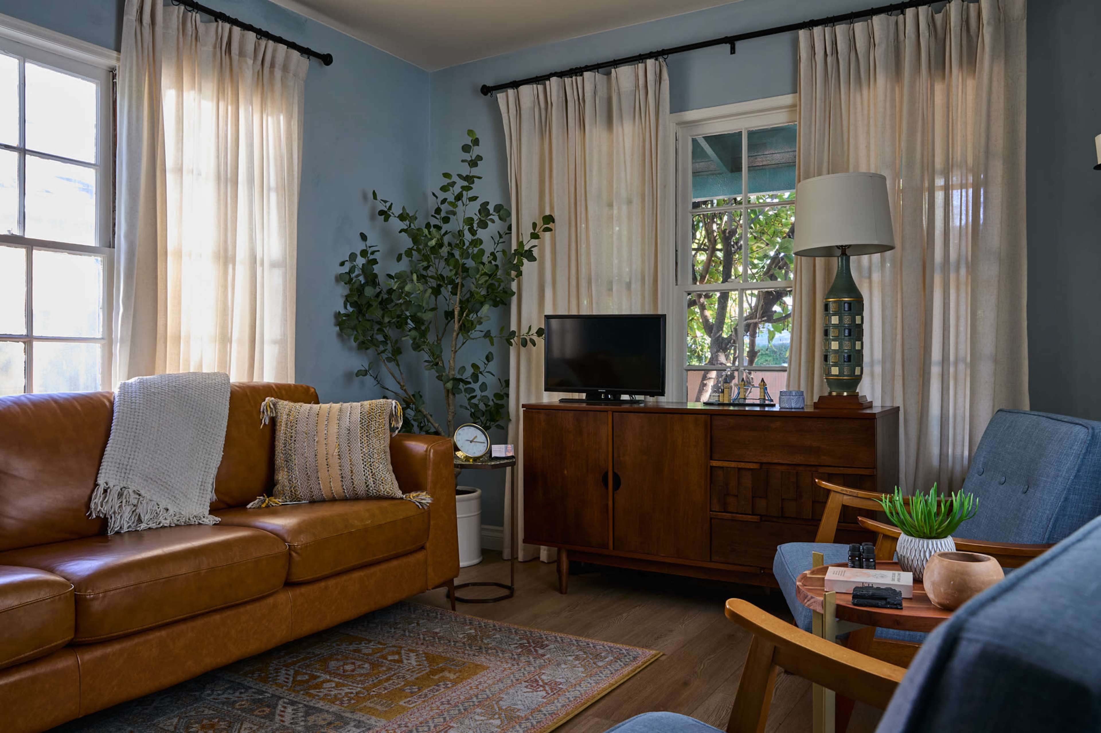 The room features a brown leather sofa, two blue chairs, a wooden cabinet with a television, and large windows dressed with light curtains.