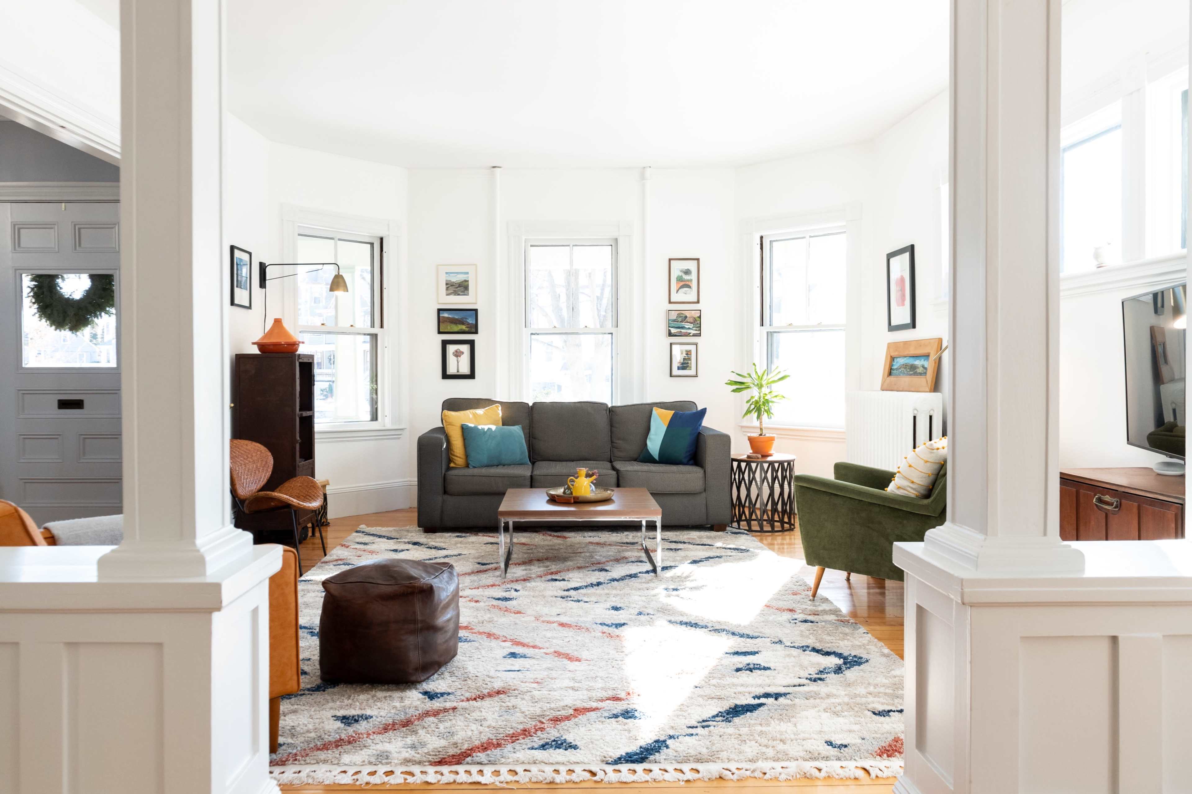 A bright living room features a gray sofa, green armchair, and a coffee table on a patterned rug, with artwork on the walls and large windows allowing natural light.