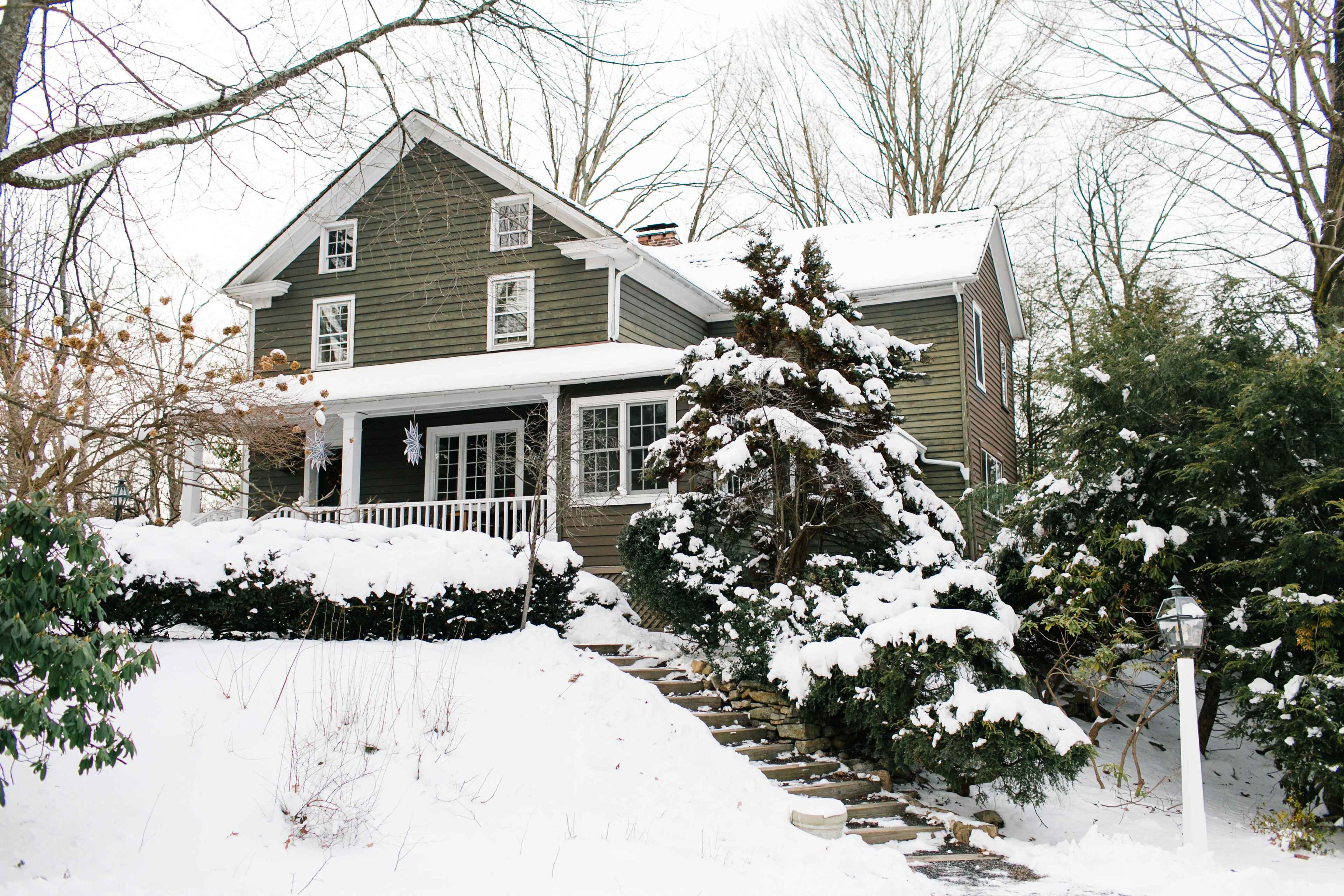 A large, two-story house with a green exterior and a front porch is surrounded by snow-covered trees and shrubs.