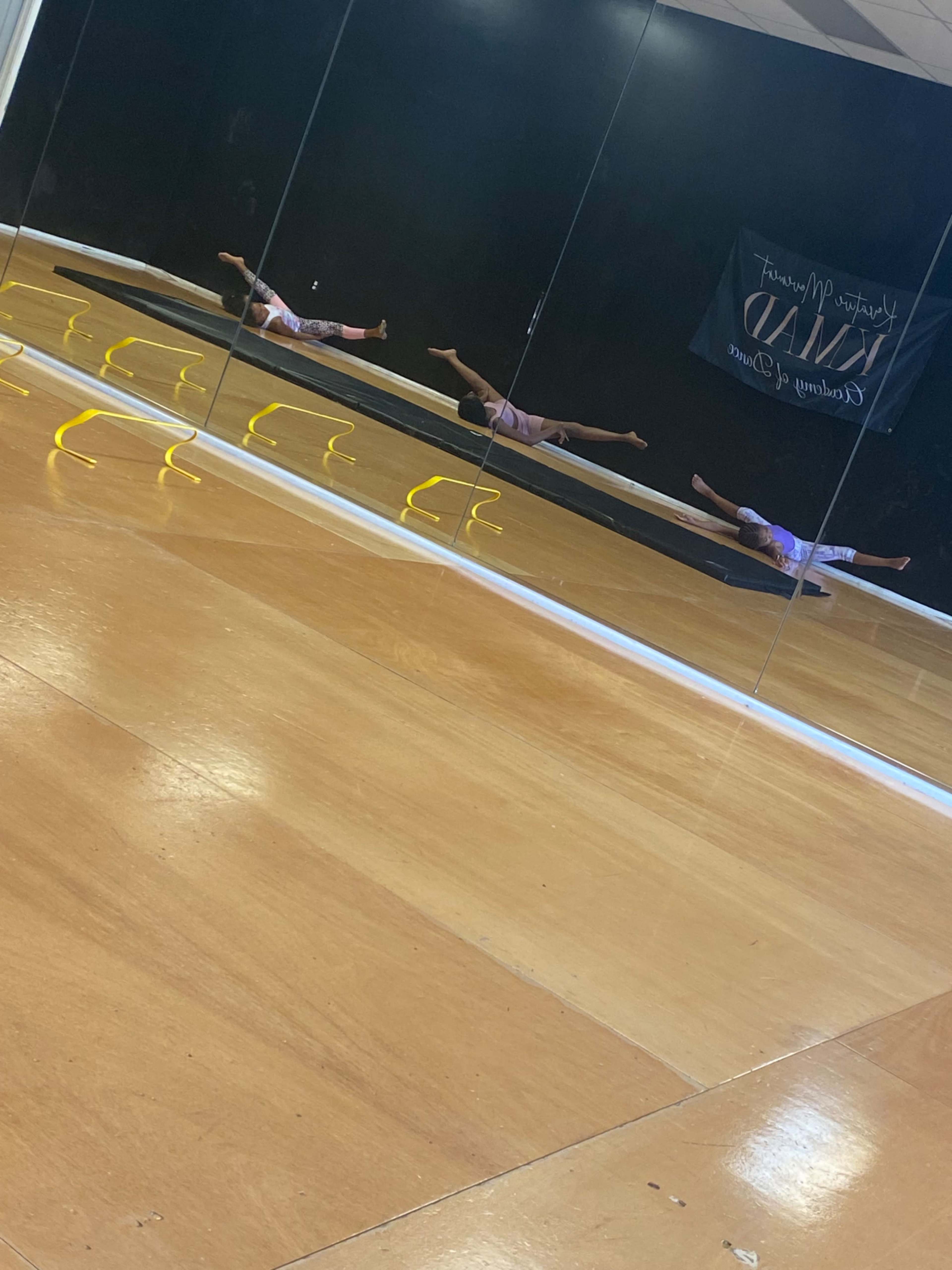 A dancer performs a leap across a mirrored studio with wooden flooring and a banner in the background.