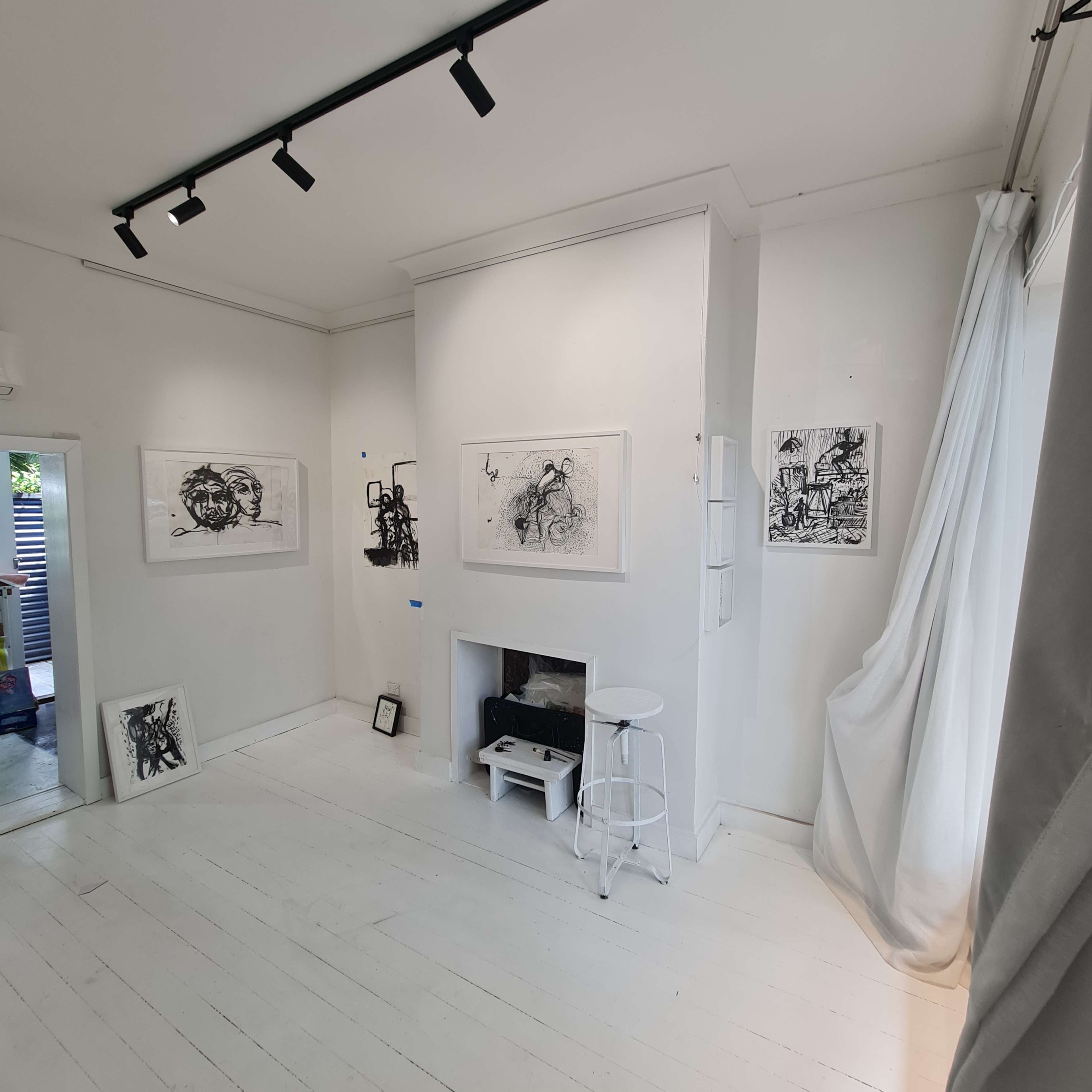 A minimalist art gallery with white walls displaying various black and white artworks, a small fireplace, and a stool in the corner.