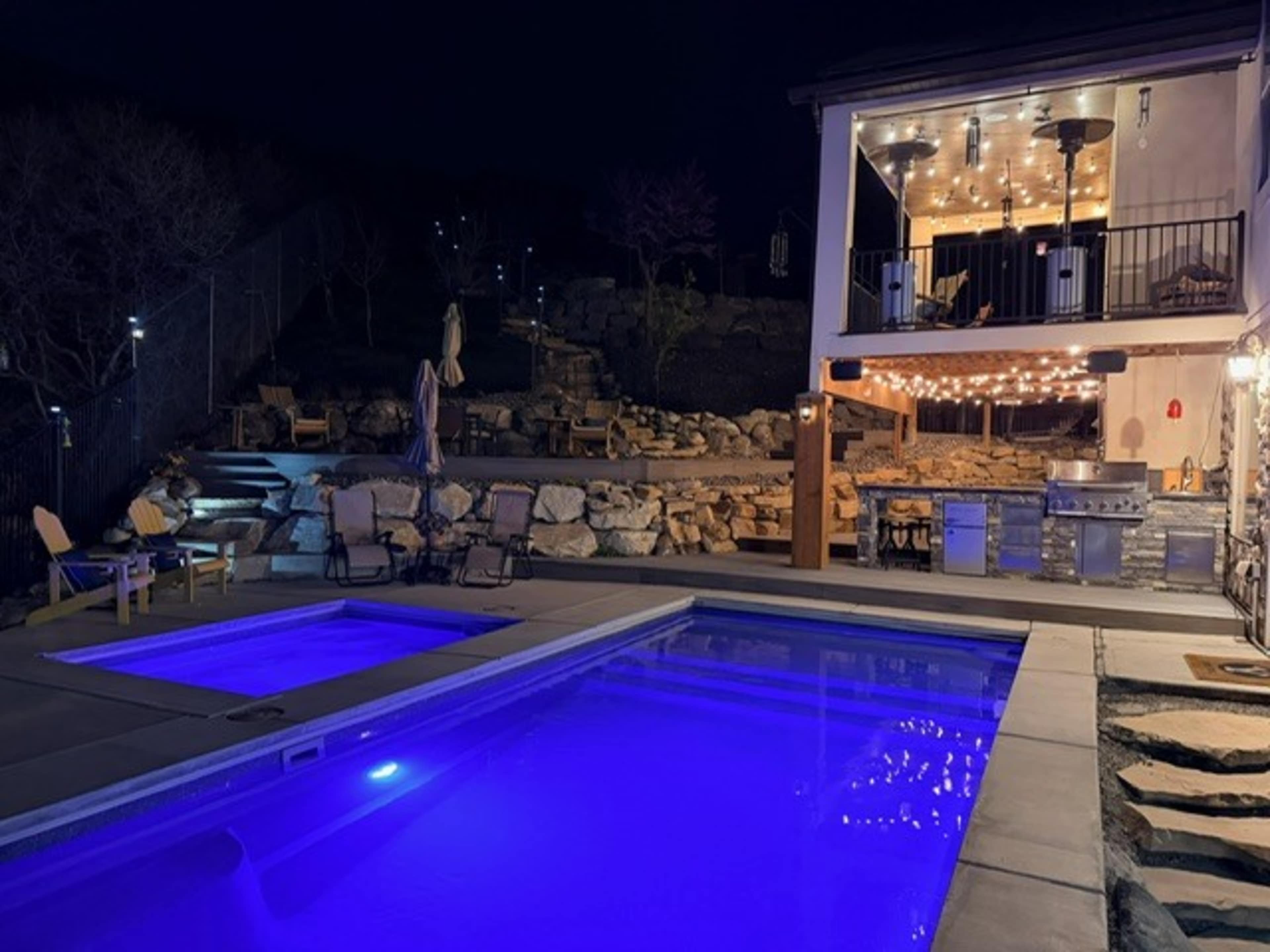 A well-lit outdoor pool area features a large pool with purple lighting, a hot tub, stone landscaping, and a two-story patio with outdoor seating and a grill.