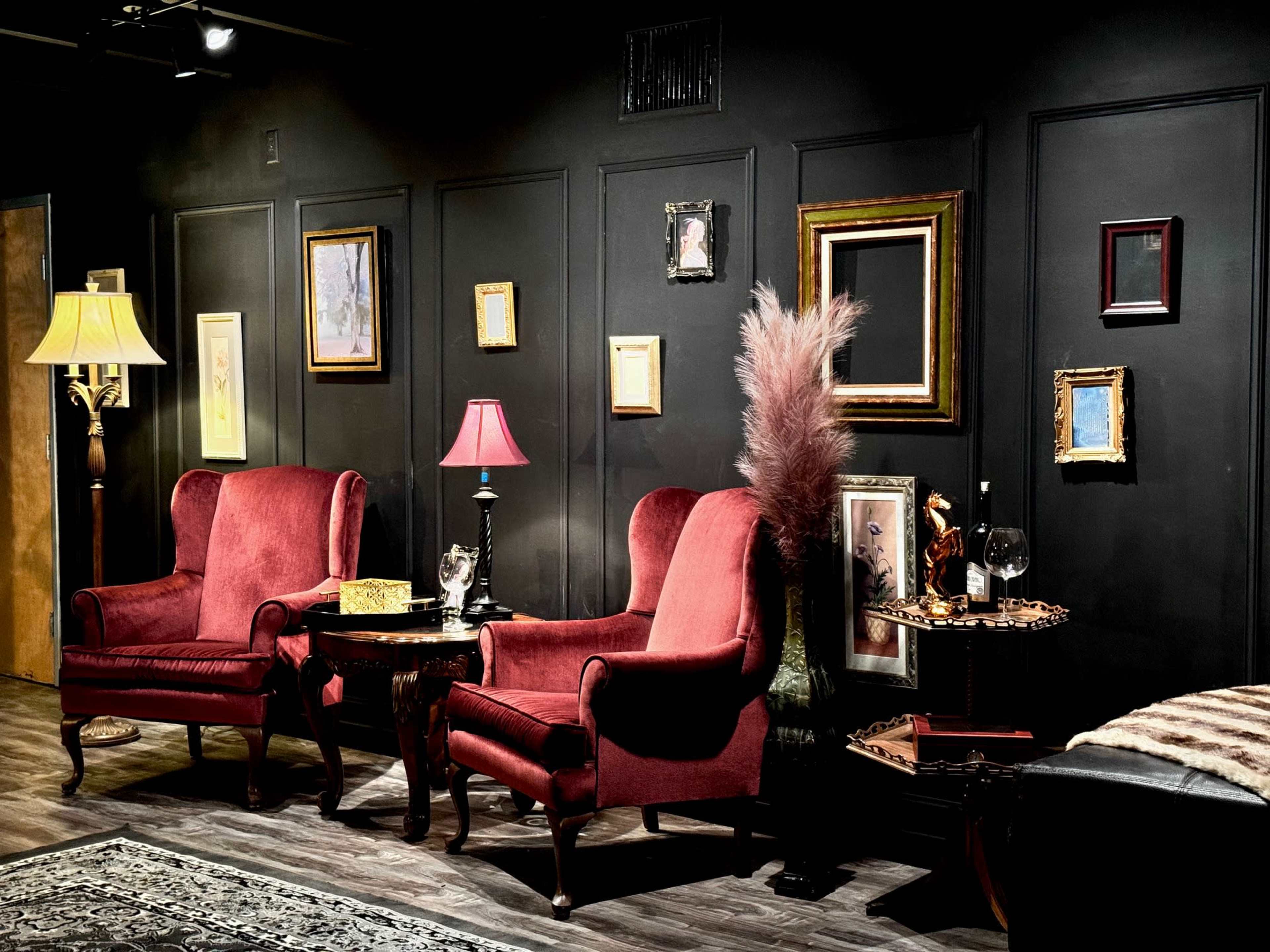 The scene features a cozy, dimly lit room with two red armchairs, a side table with a lamp and decorative items, and various framed pictures on a black wall.