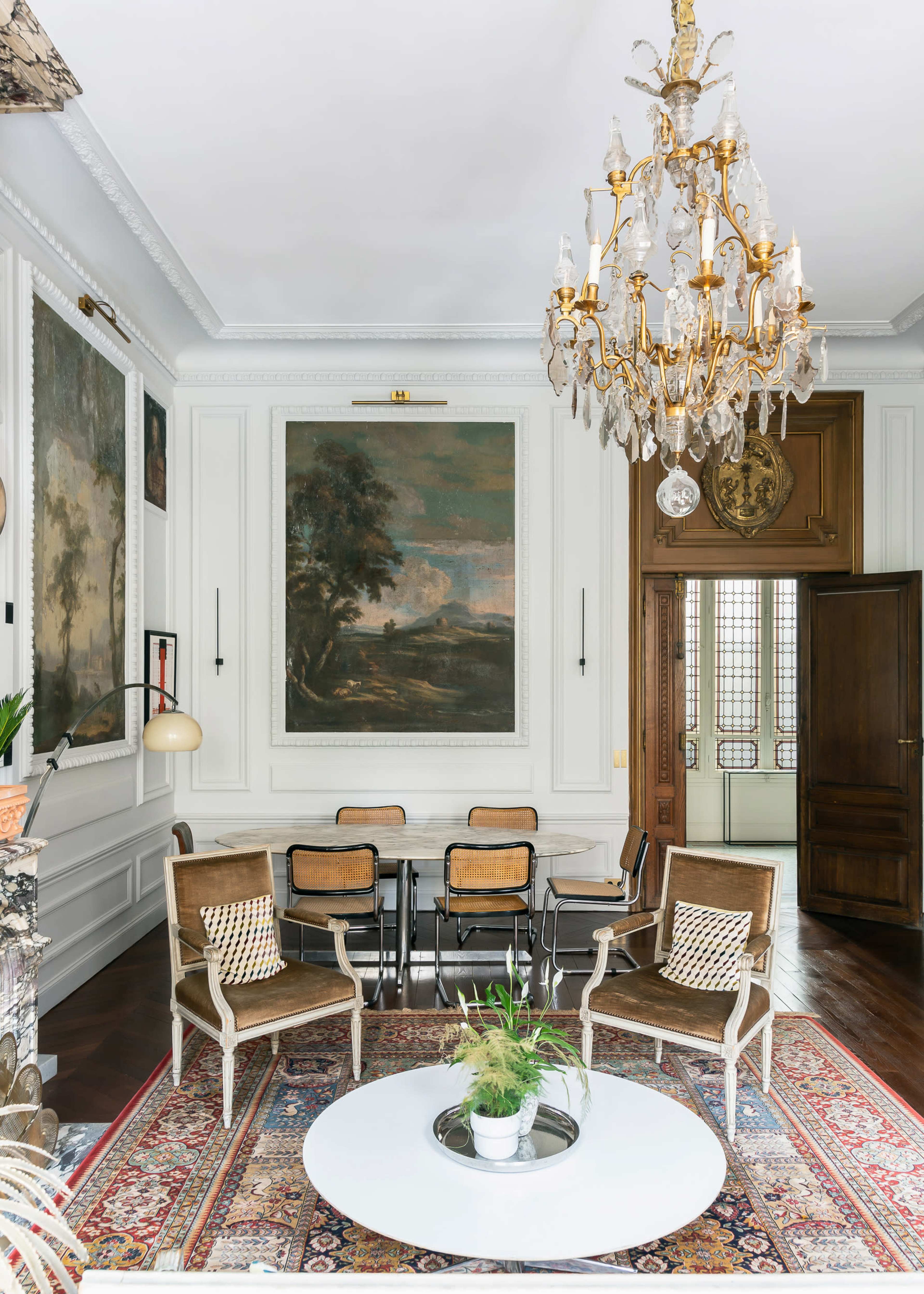 The room features a dining area with a round table surrounded by chairs, flanked by large paintings on the walls and adorned with a chandelier overhead.