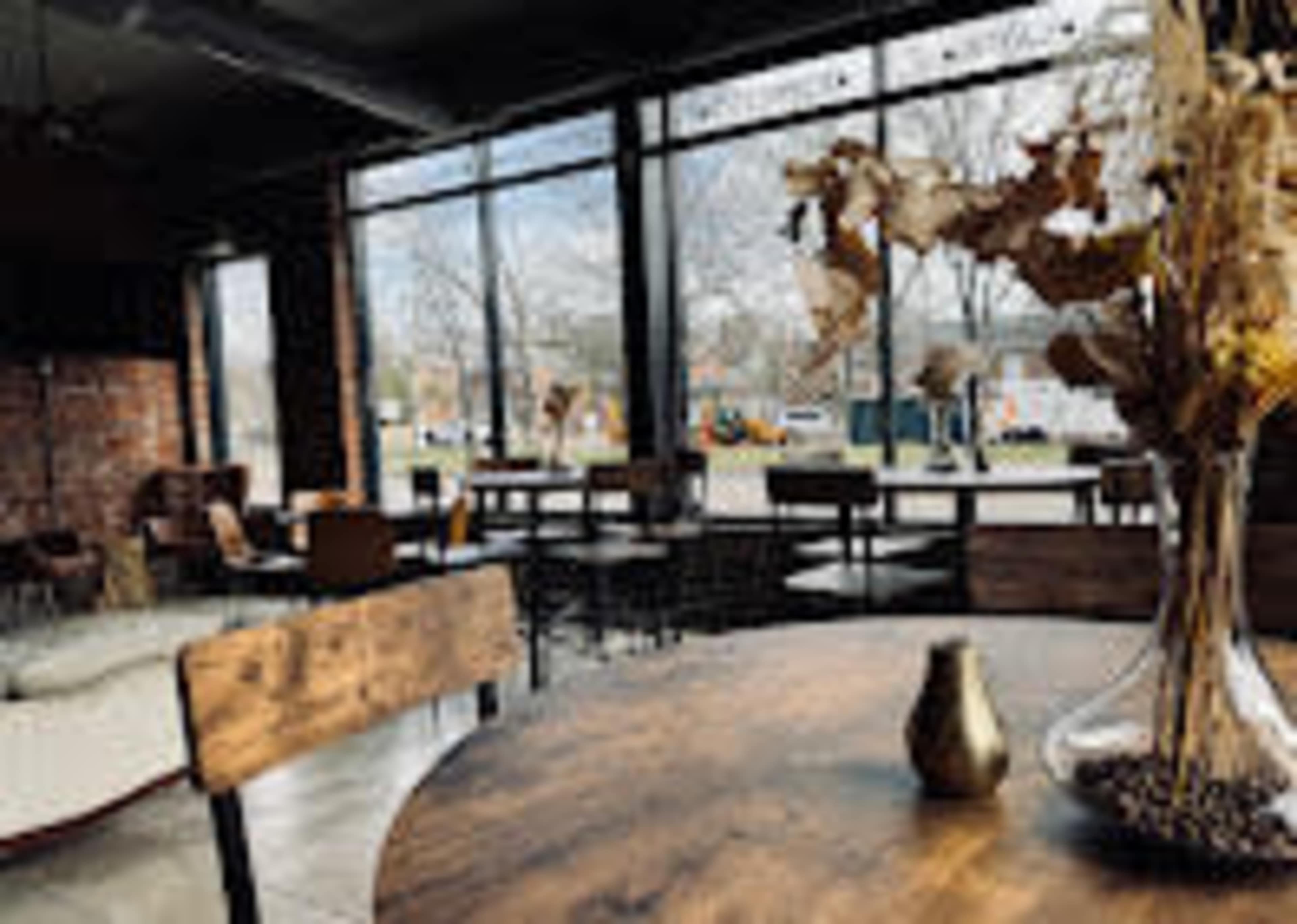 The image shows a cozy café interior with large windows, wooden tables, and a decorative vase on the table.