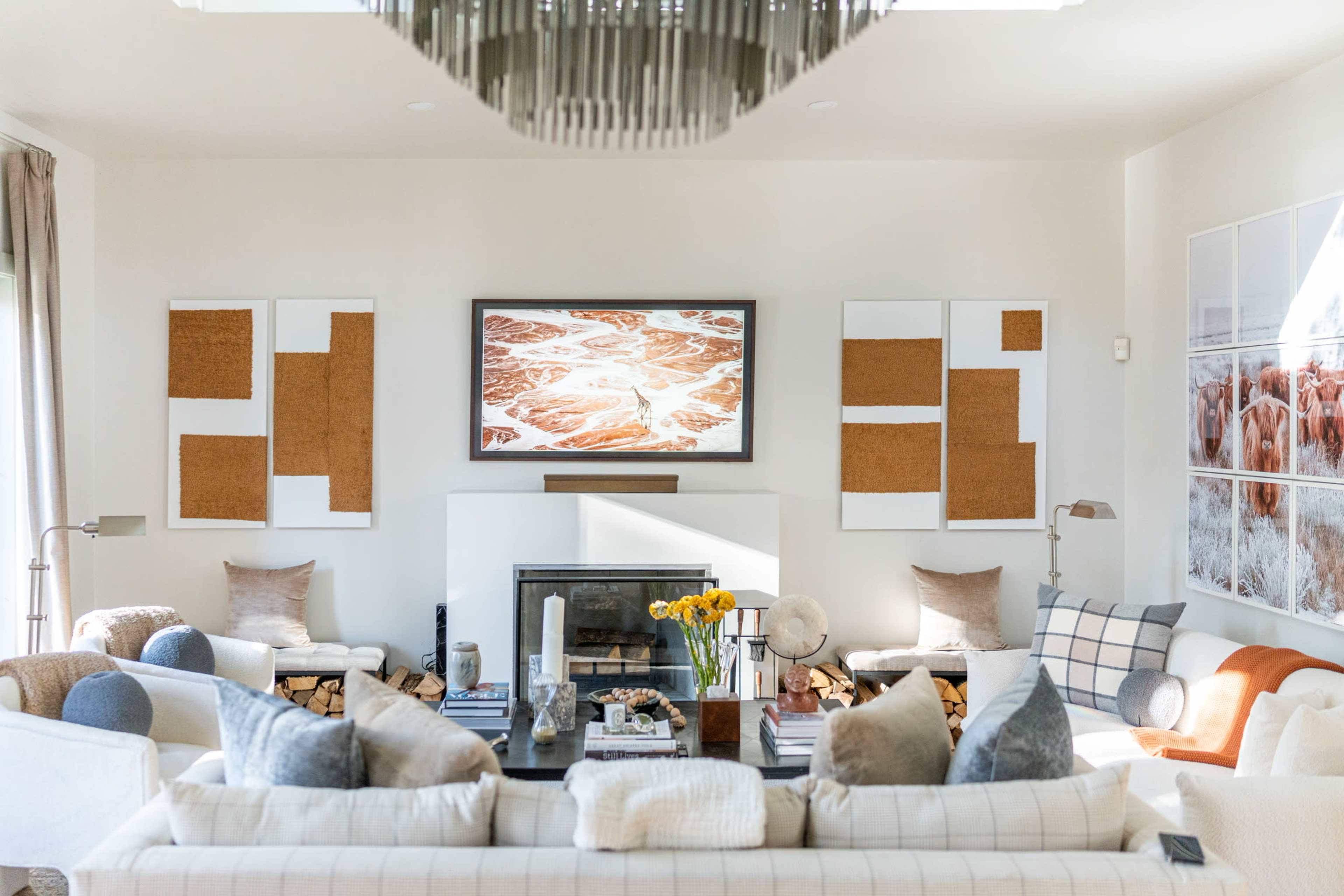 A modern living room with a large television above a fireplace, surrounded by artwork and cozy seating arrangements featuring various cushions and a coffee table.