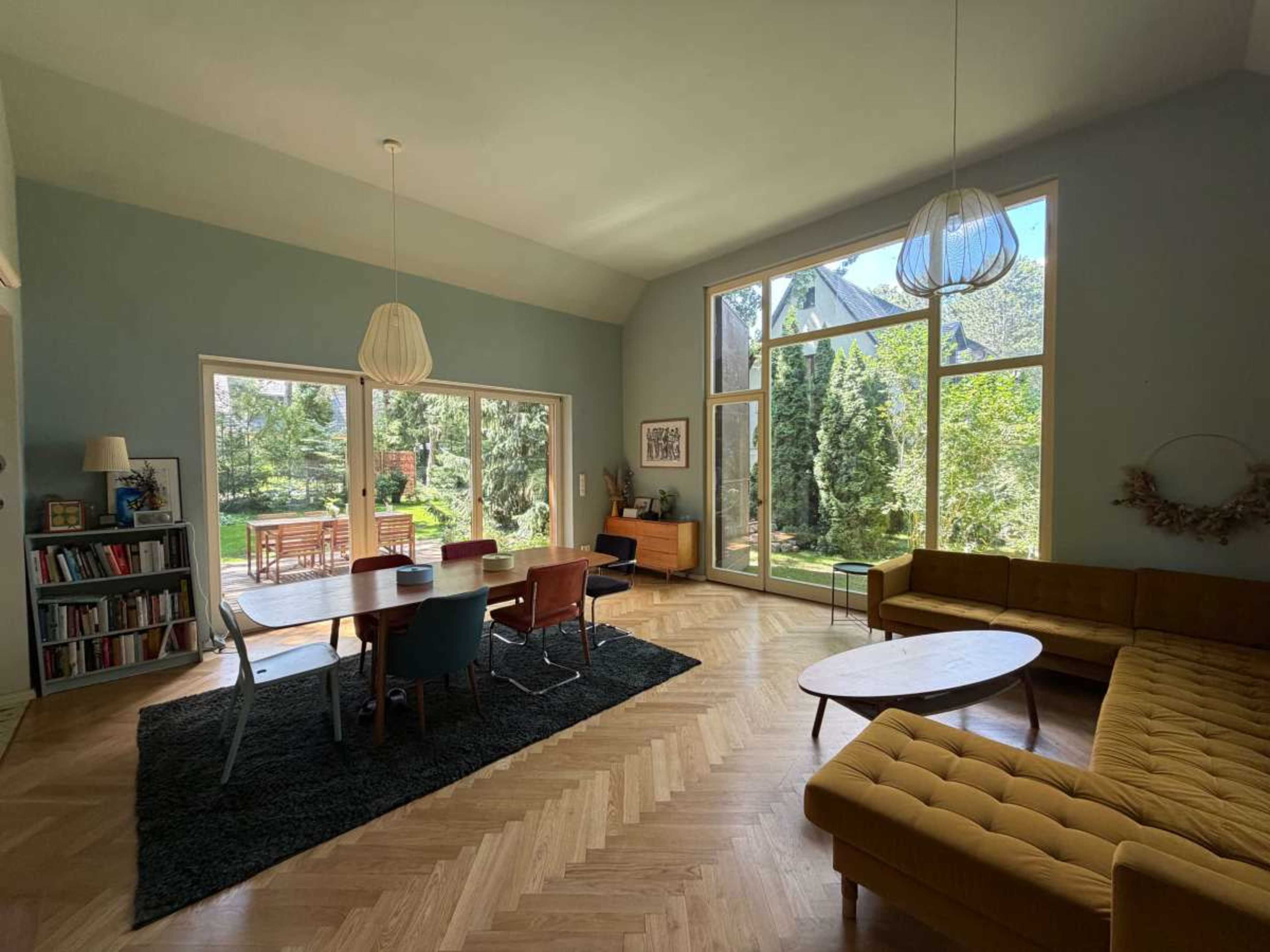 A spacious, modern living area features a large dining table surrounded by colorful chairs, a sectional sofa, and large windows overlooking a garden.