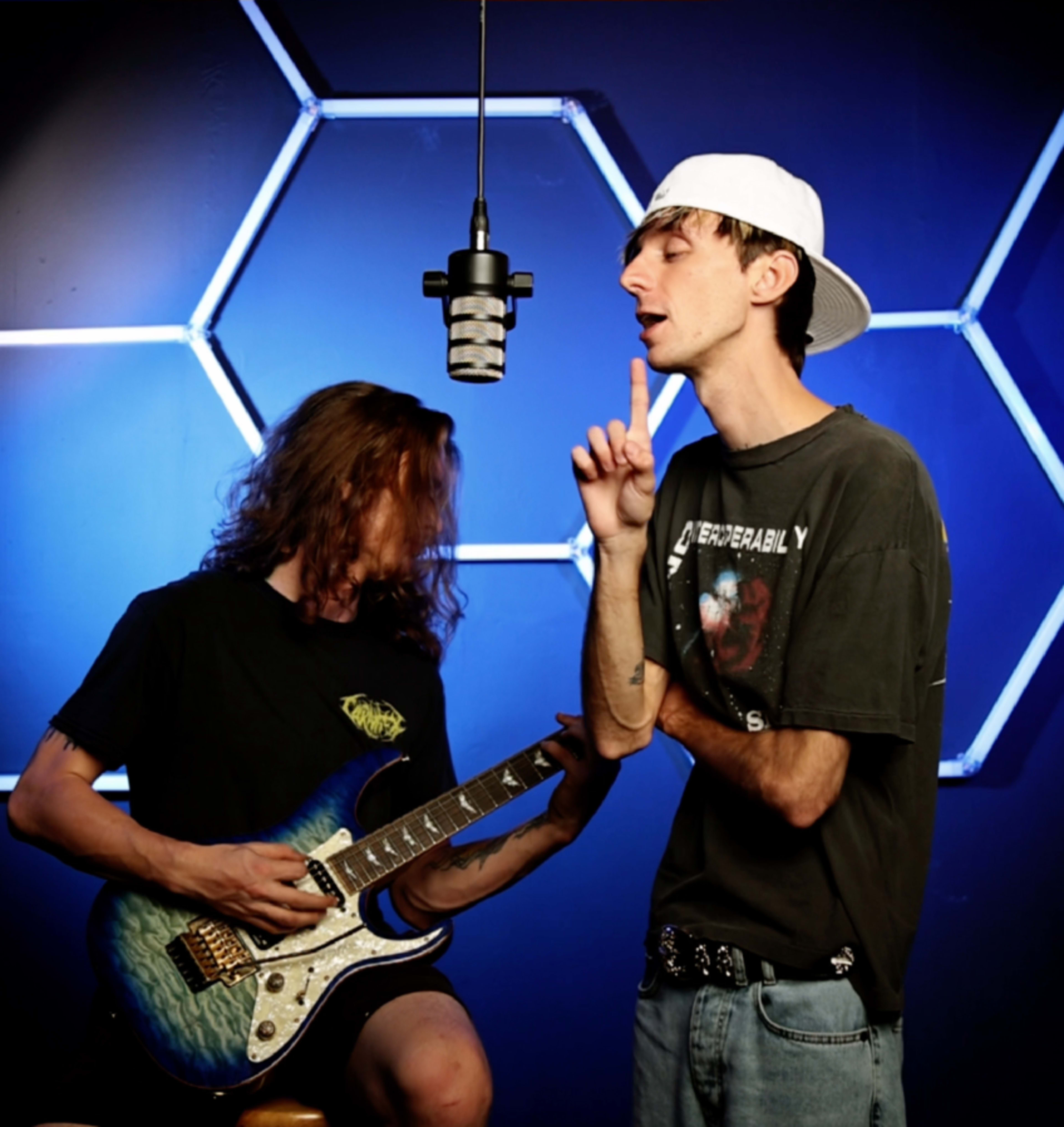 Two young men are performing together, one singing into a microphone while the other plays an electric guitar against a blue backdrop with hexagonal lights.