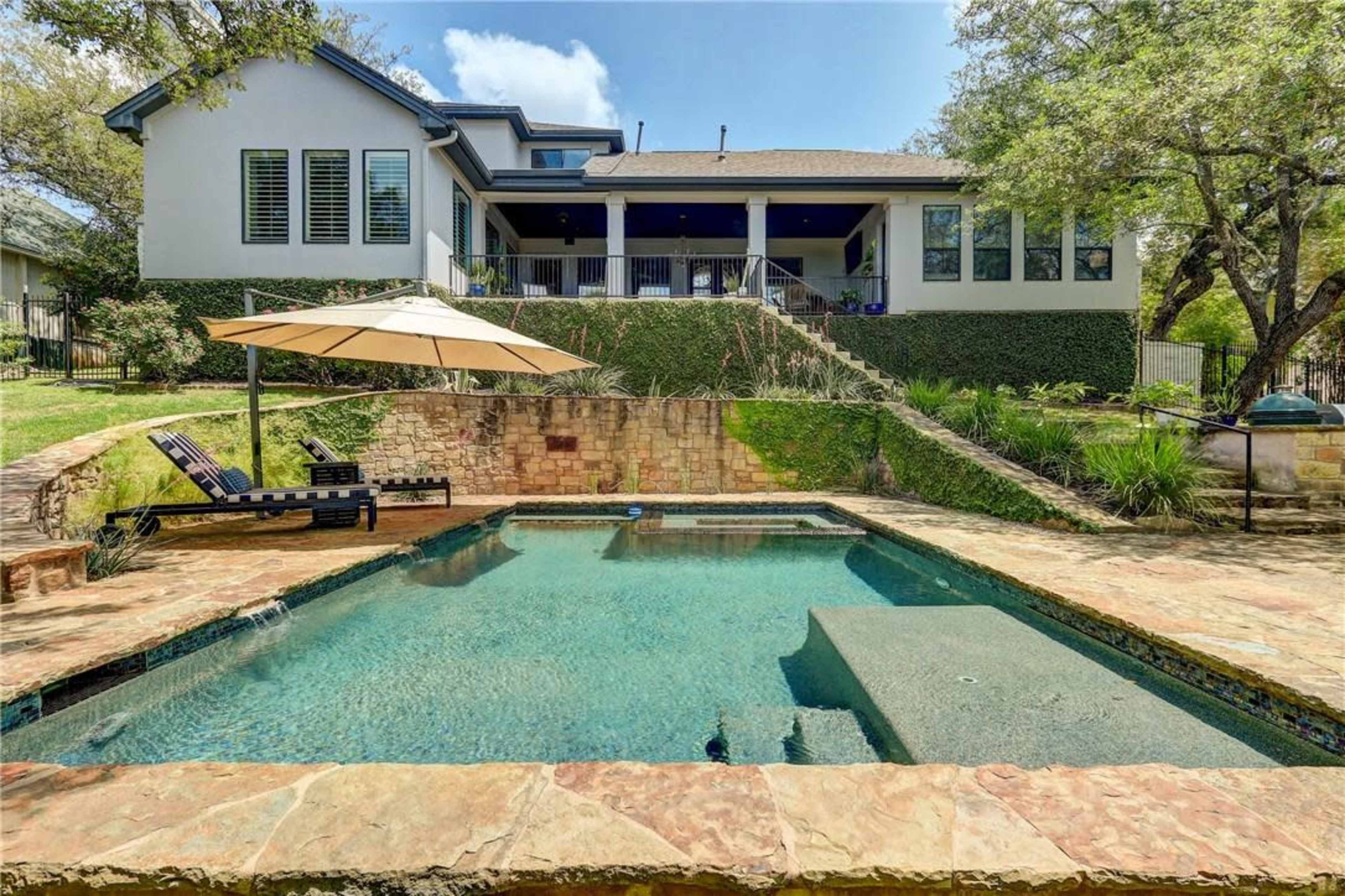 A backyard features a swimming pool surrounded by stone edging and a patio with lounge chairs and an umbrella, adjacent to a two-story house.