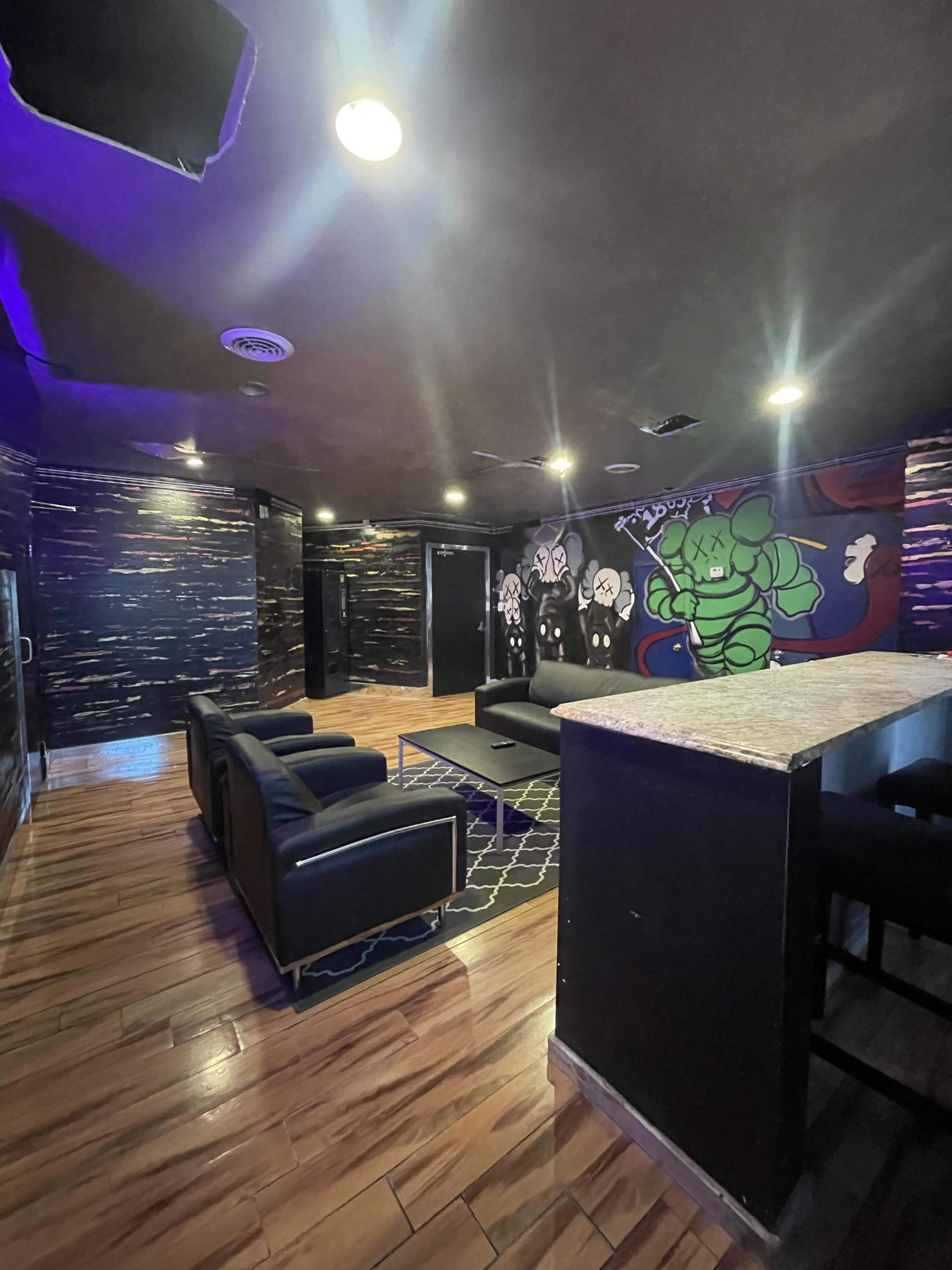 The image shows a dimly lit lounge area with black sofas, a coffee table, and a bar top, featuring colorful wall murals.