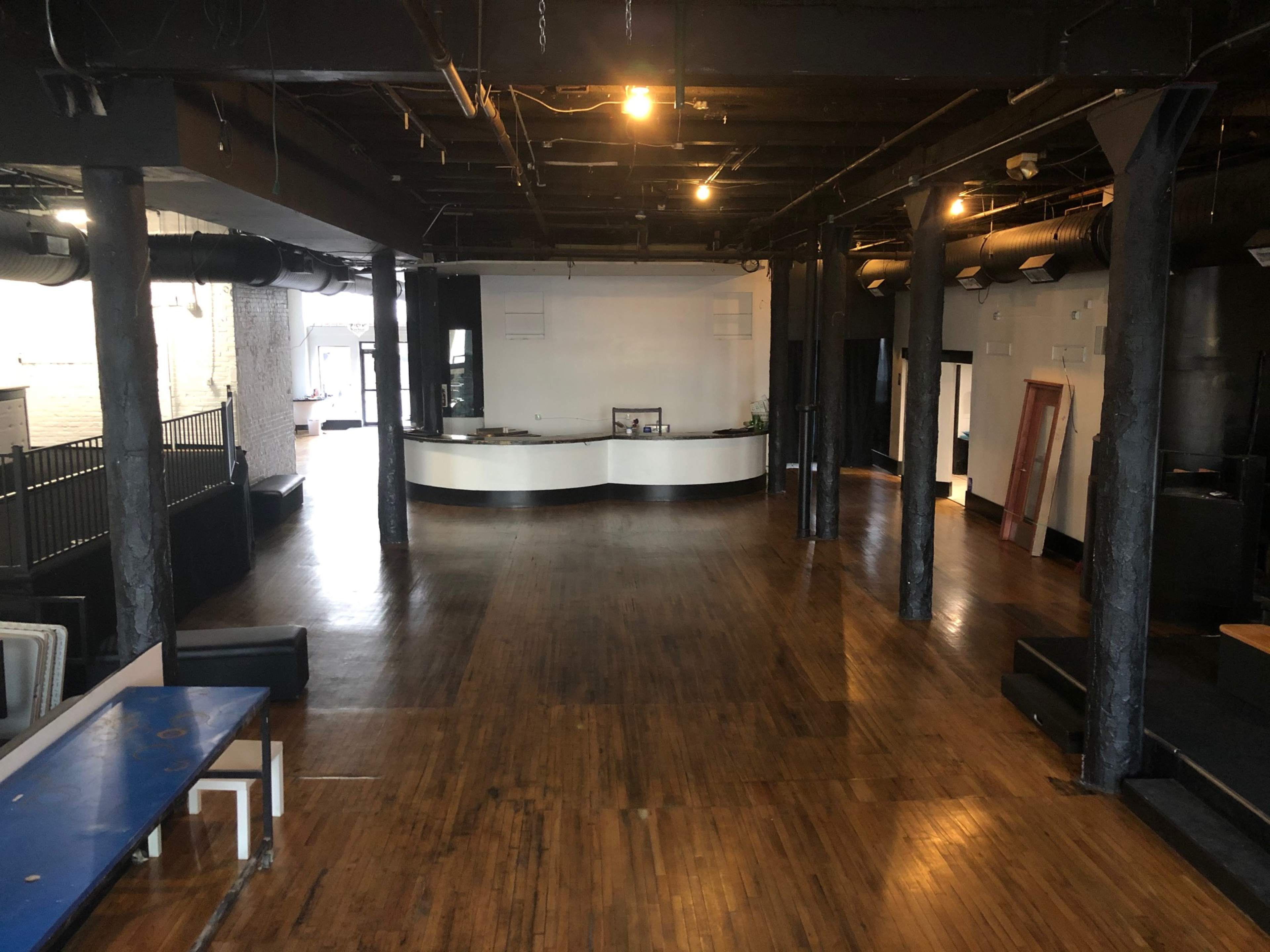 The image shows a spacious, empty interior of a venue featuring dark wooden floors, tall pillars, and a reception area with a curved counter.