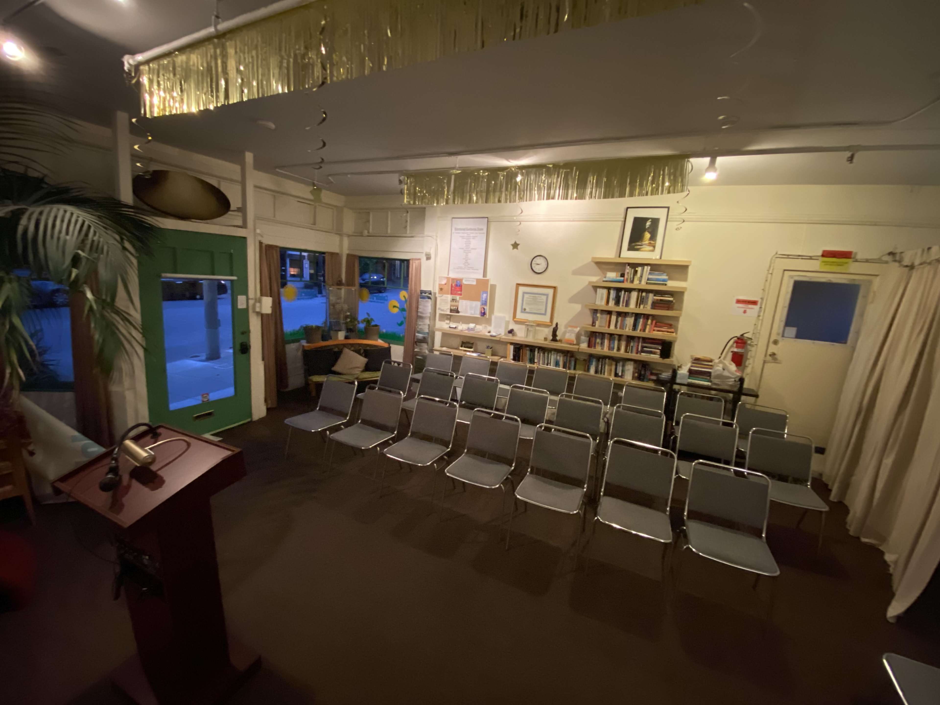The image shows an empty room set up with metal chairs facing a wooden podium, with bookshelves and a green door in the background.