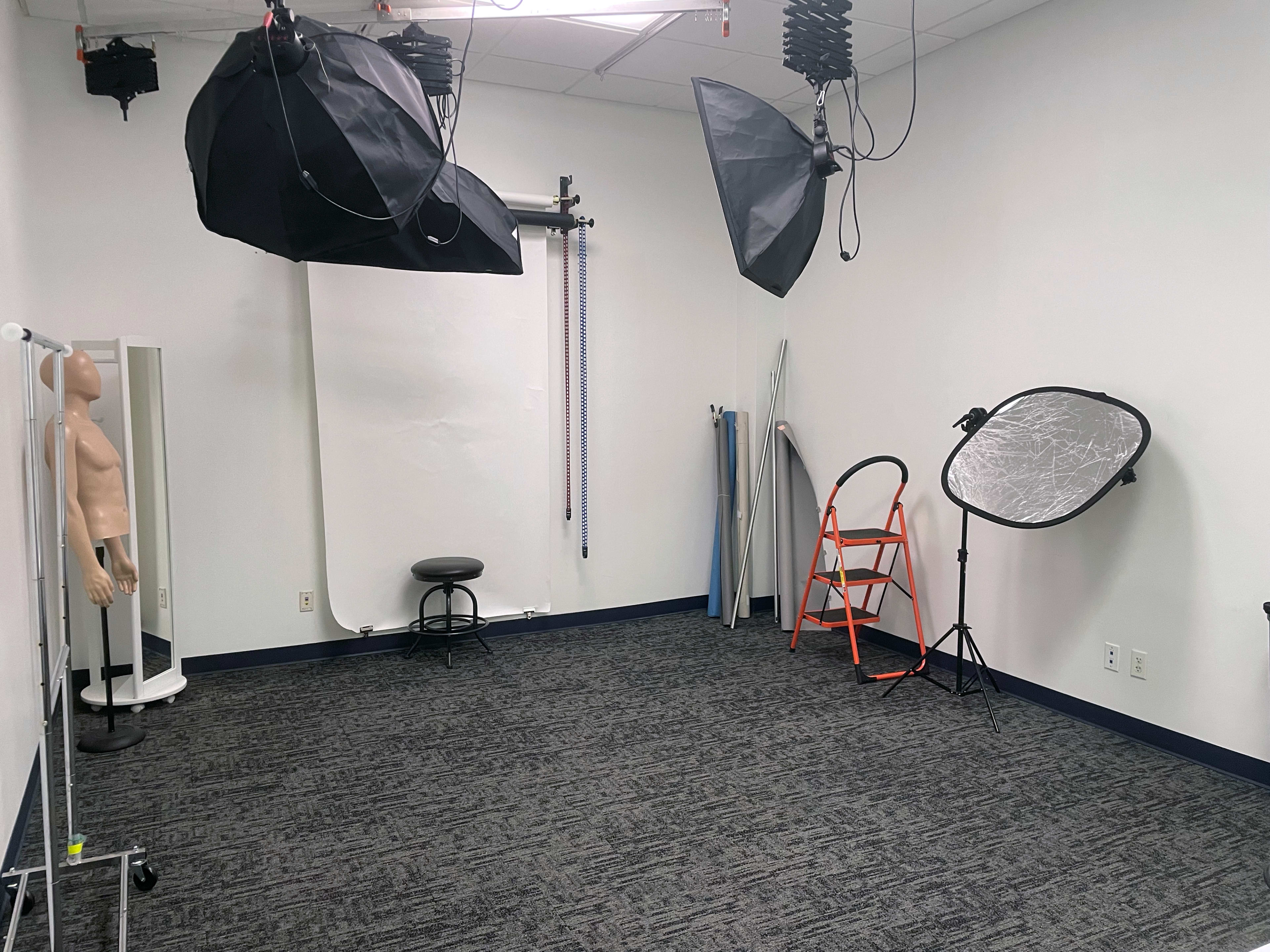 A photography studio equipped with lighting setups, a mannequin, a stool, a ladder, and a backdrop.