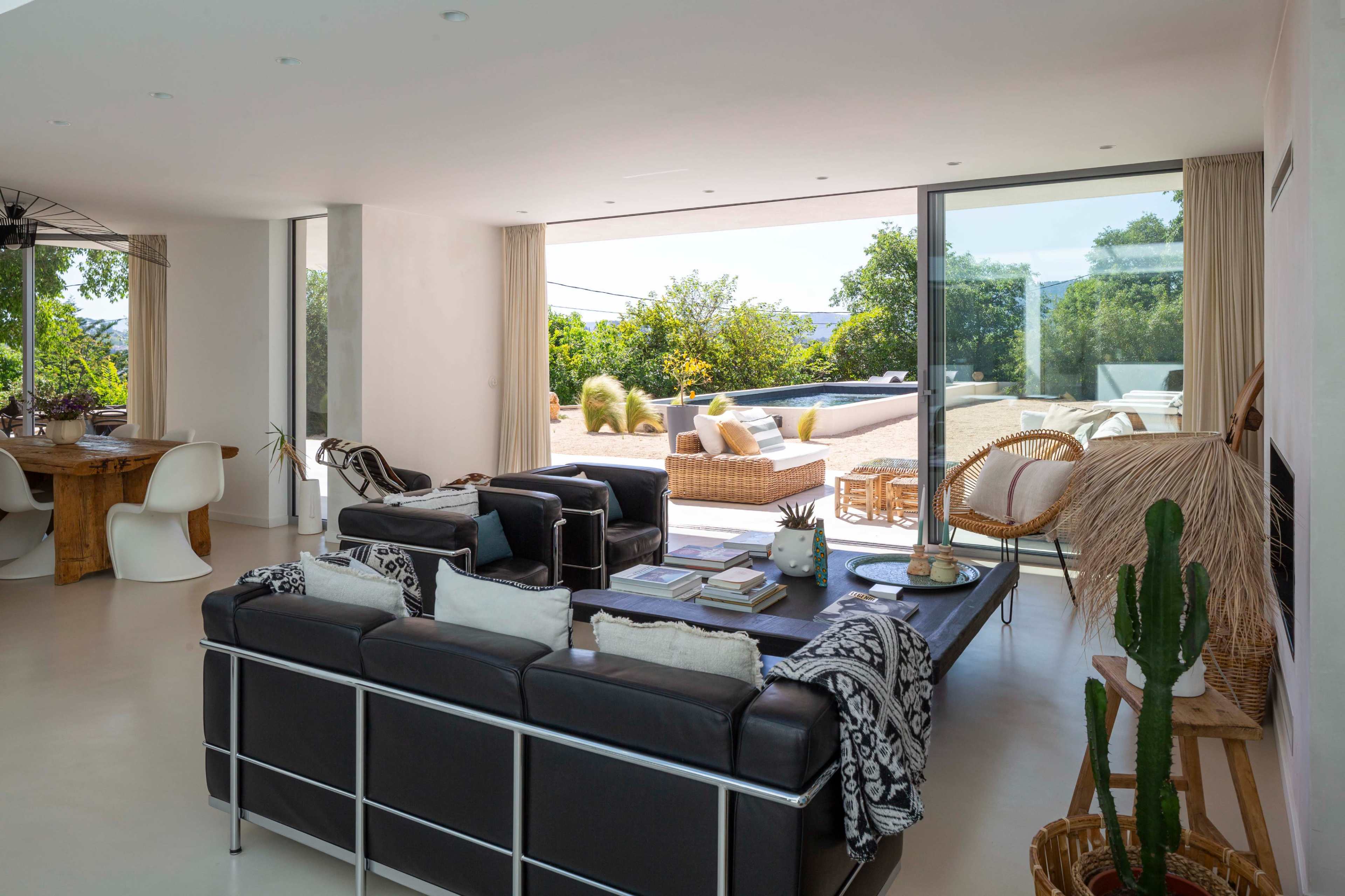 The image shows a modern living room with large glass doors leading to an outdoor patio, featuring a mix of contemporary furniture and natural decor elements.