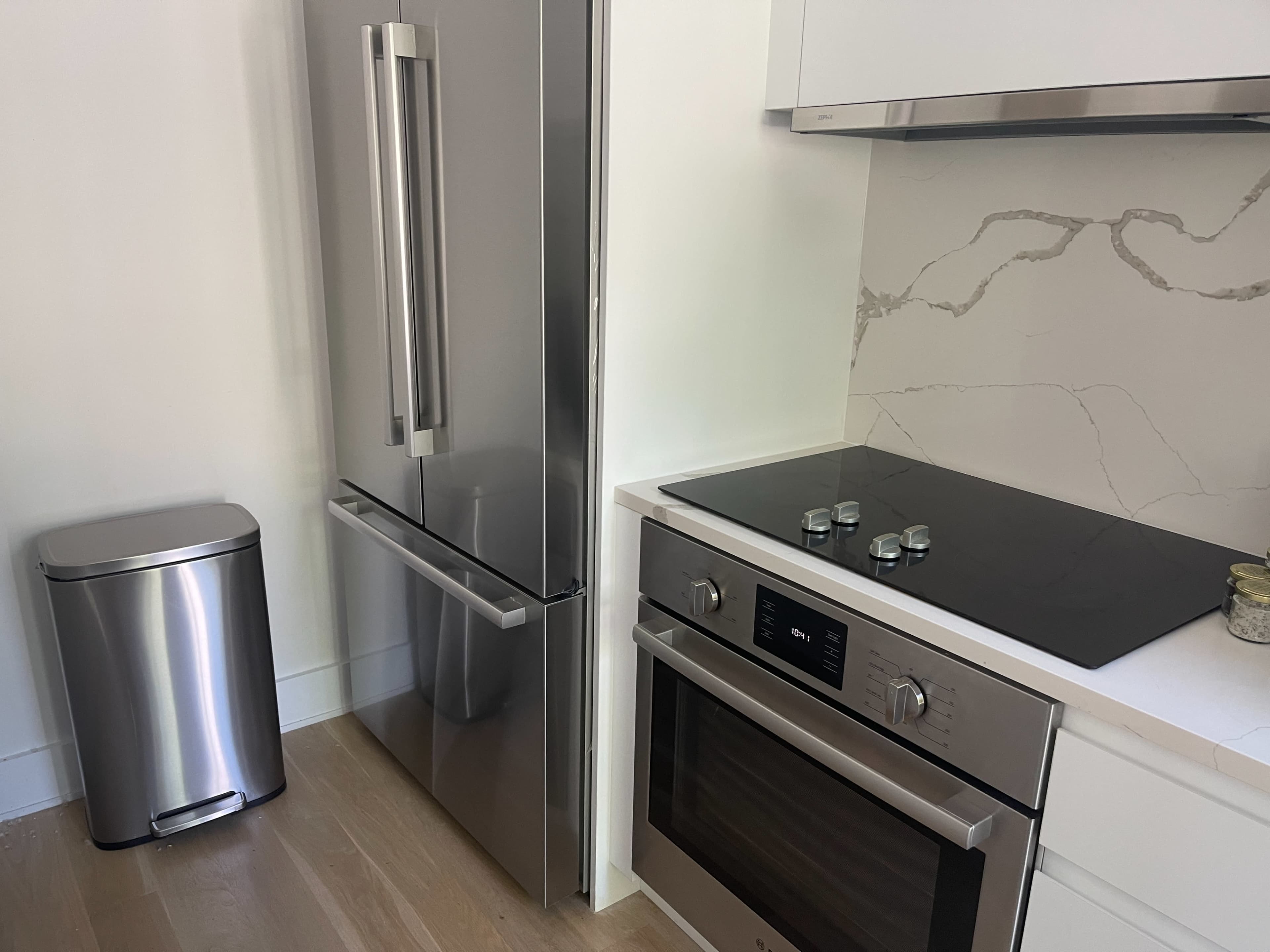 The image shows a modern kitchen corner featuring a stainless steel refrigerator, a built-in oven and cooktop, and a garbage can.