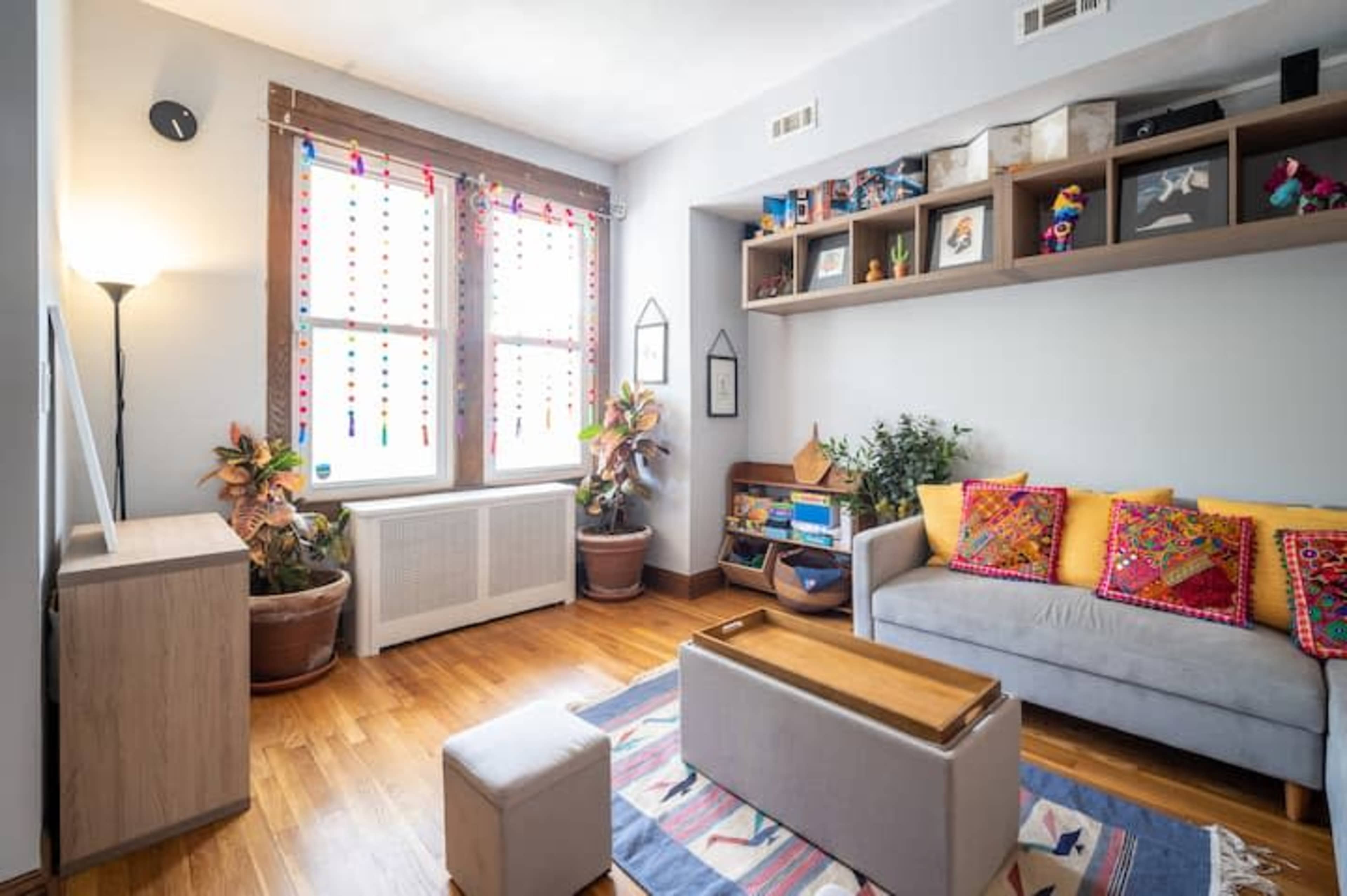 A bright living room featuring a gray sofa adorned with colorful pillows, a wooden coffee table, and shelves displaying various items, with large windows allowing natural light to enter.