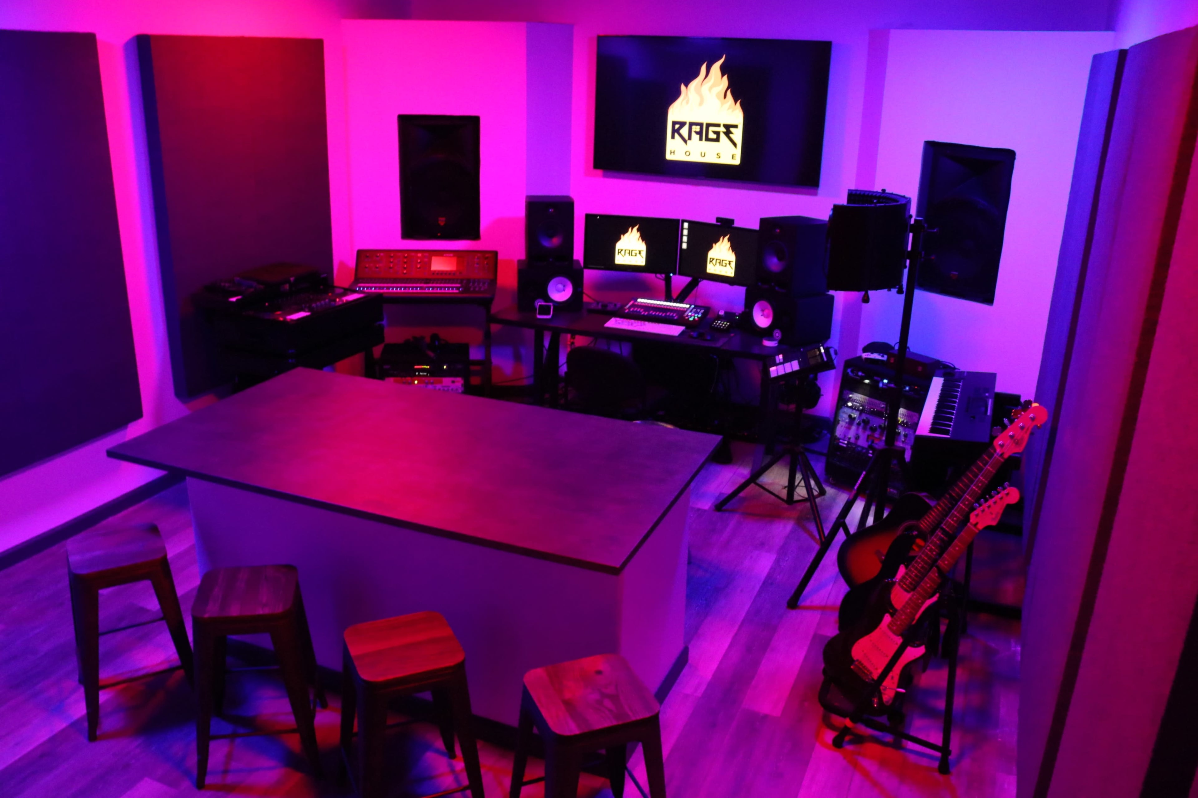 A music production studio featuring a large central table, several audio equipment pieces, and soundproofing panels on the walls, illuminated by purple and pink lighting.